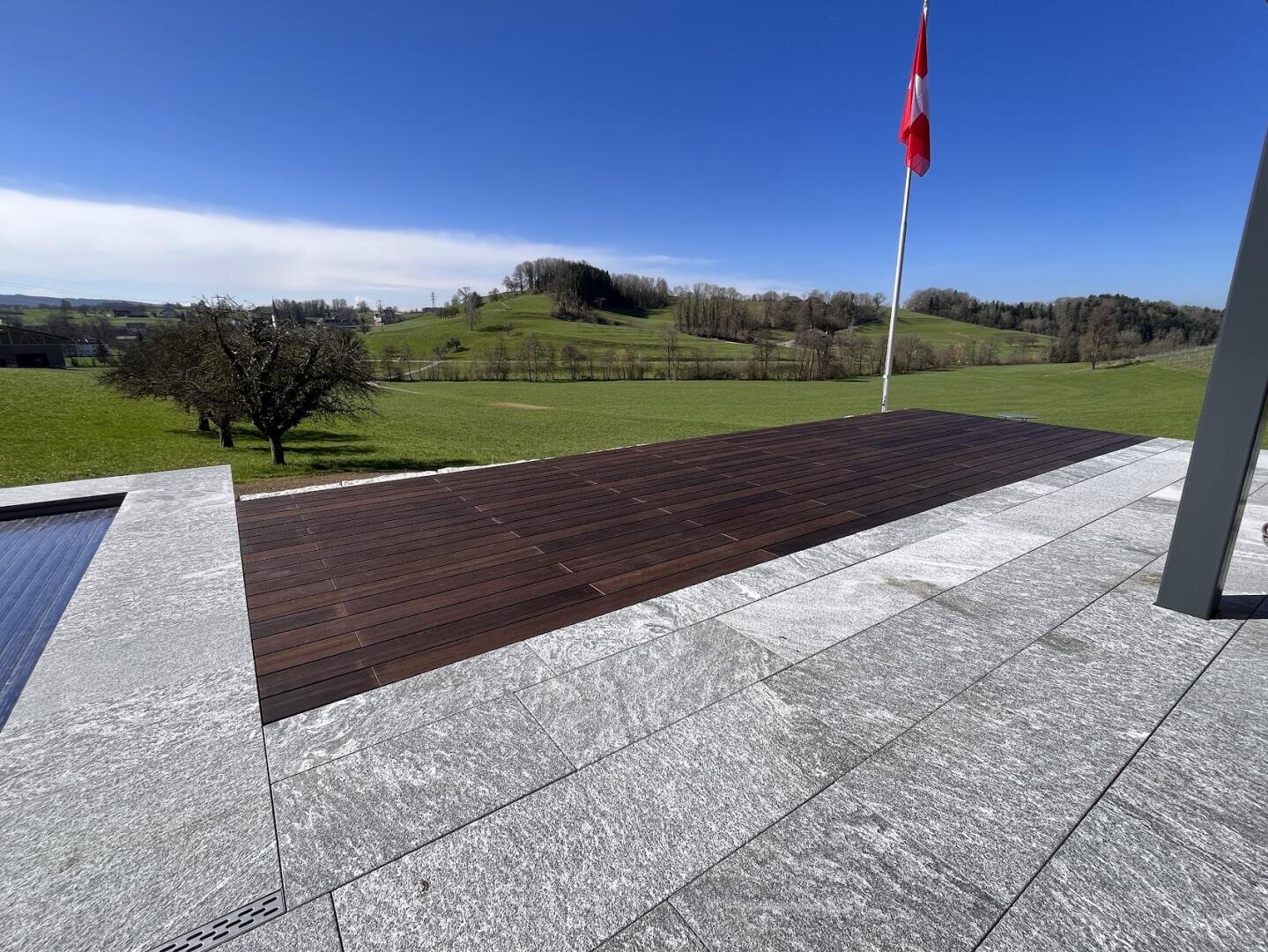 Eine steinerne Terrasse mit einem hölzernen Deckteil blickt auf grüne Grashügel unter einem klaren blauen Himmel. Rechts steht eine Schweizer Flagge an einem Fahnenmast, und ein paar Bäume säumen die Landschaft.