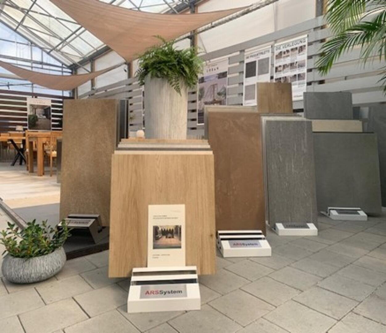 Auslage mit verschiedenen großen Fliesenmustern in neutralen Tönen, aufrecht in einem Ausstellungsraum angeordnet, mit Informationsschildern, Pflanzen und Postern im Hintergrund. Das Licht fällt durch eine Glasdecke darüber ein.
