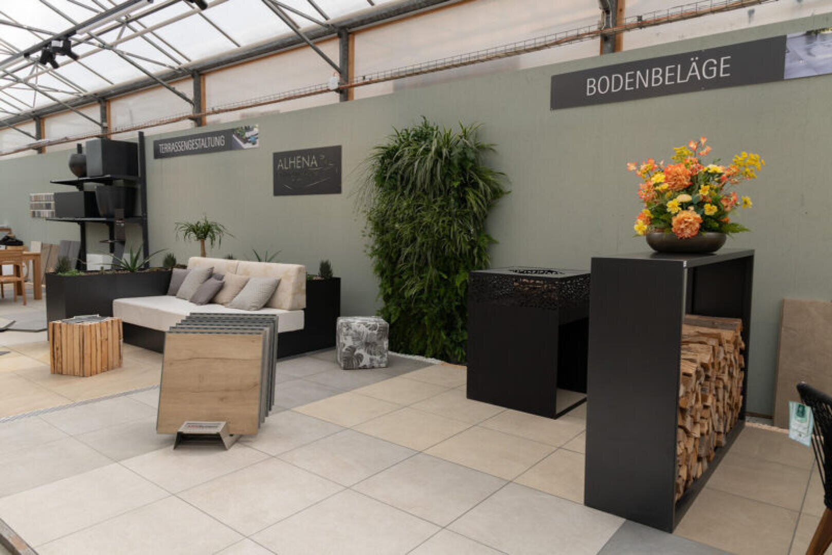 In einem modernen Ausstellungsraum werden Gartenmöbel, Topfpflanzen, ein Sofa und Bodenbelagsmuster ausgestellt. An der Wand hängen Schilder mit der Aufschrift Bodenbeläge und Terrassenbelegung. Brennholz und ein Blumenarrangement stehen auf schwarzen Ständern.