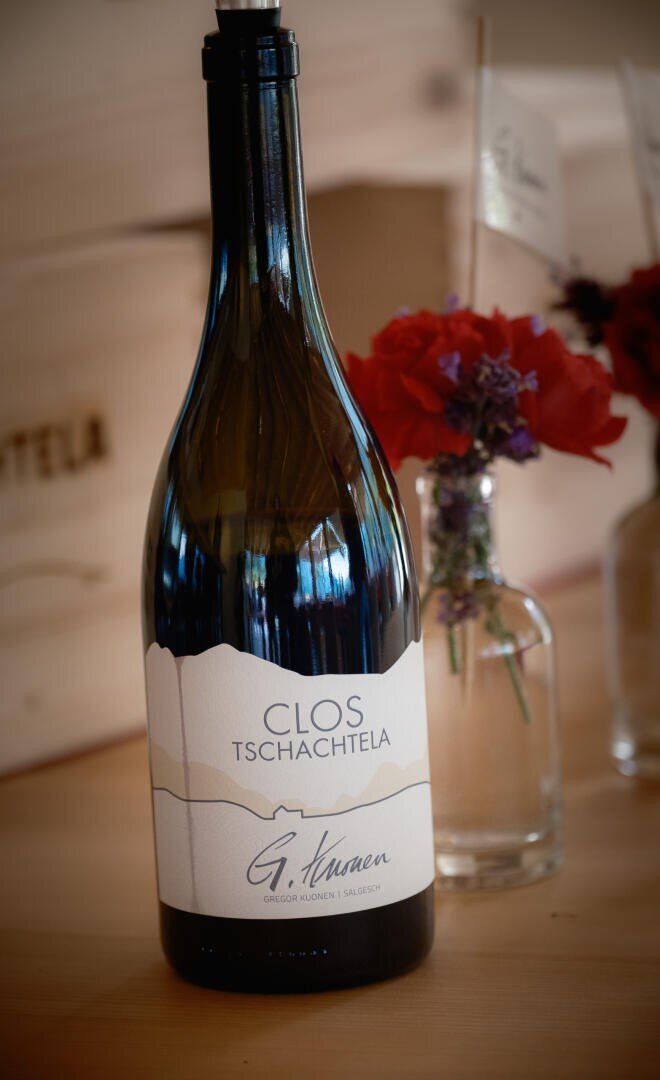 Eine Weinflasche mit der Aufschrift "Clos Tschachtetla" steht auf einer hölzernen Unterlage, während im unscharfen Hintergrund kleine Glasvasen mit roten Blumen zu sehen sind.