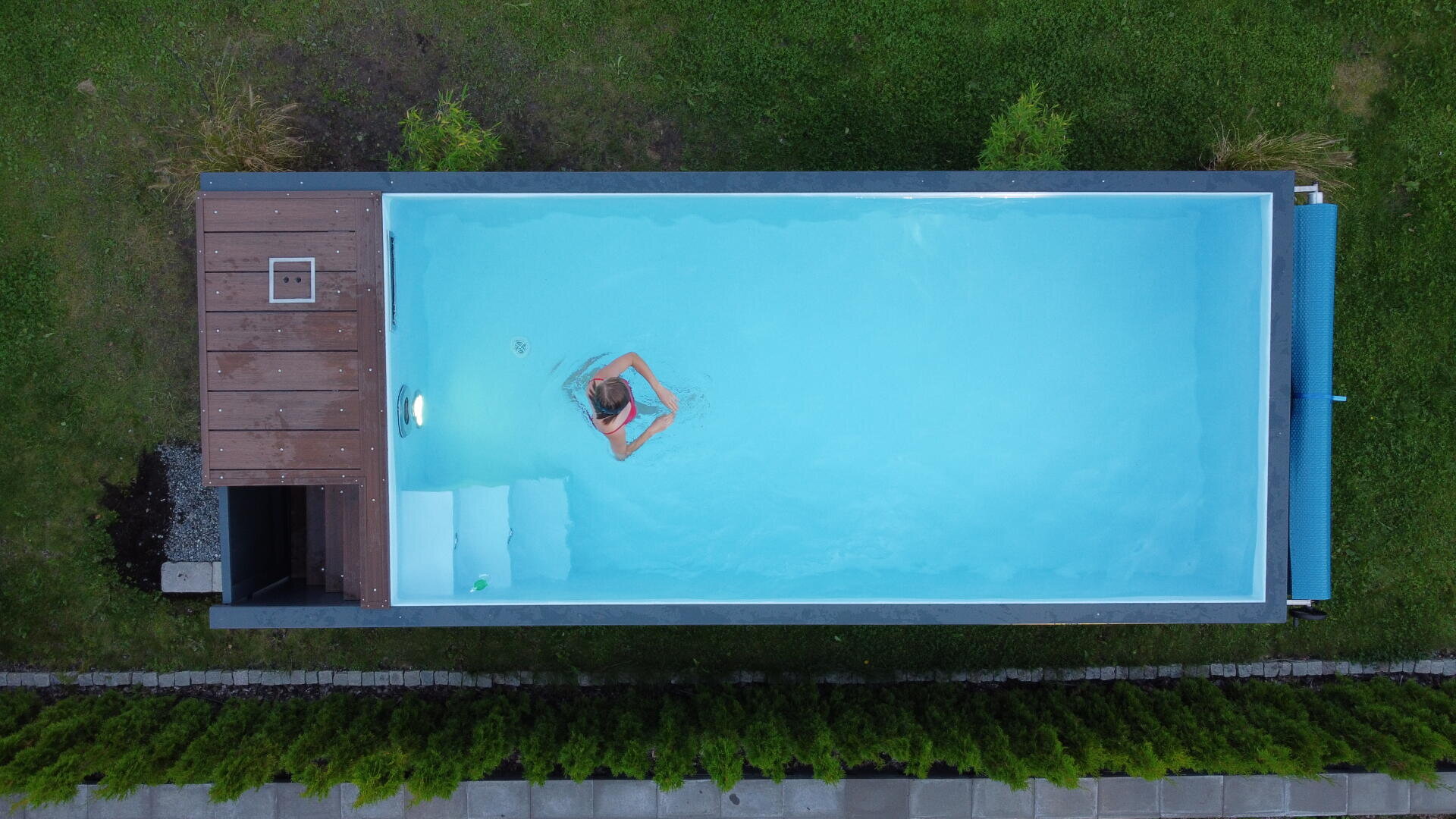 A person swims alone in a rectangular outdoor pool surrounded by grass and shrubs, viewed from above. The pool has a wooden deck on one end and clear blue water.