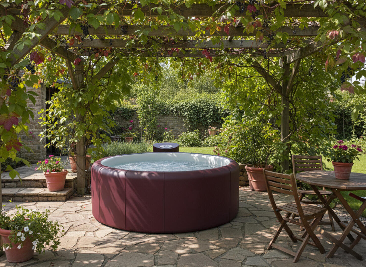Ein runder, weinroter Whirlpool steht auf einer Steinterrasse unter einer begrünten Pergola, umgeben von Topfpflanzen. In der Nähe steht ein Holztisch mit Stühlen im Schatten, während im Hintergrund ein üppiger Garten zu sehen ist.