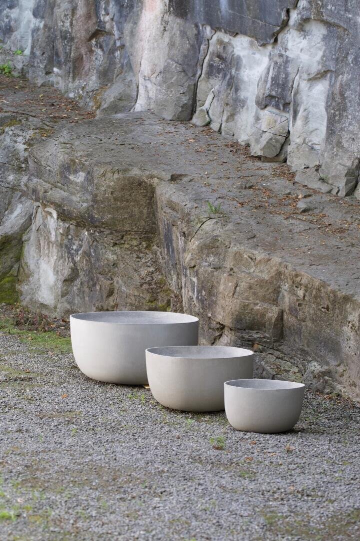 Three round, white, minimalist planters of varying sizes are placed in a row on a gravel surface, next to a rugged, rocky stone wall.