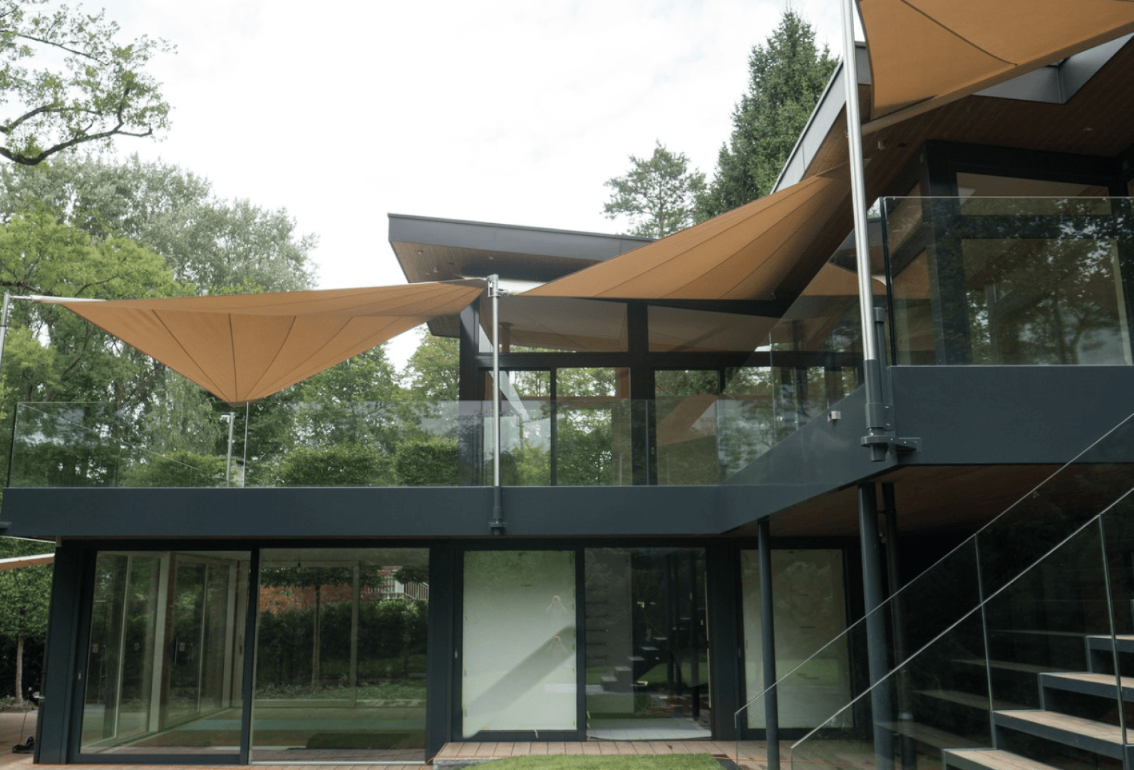 Modern two-storey house with large glass windows and doors, with beige triangular awnings over the terrace. Surrounded by greenery and trees, with metal railings and a visible external staircase.