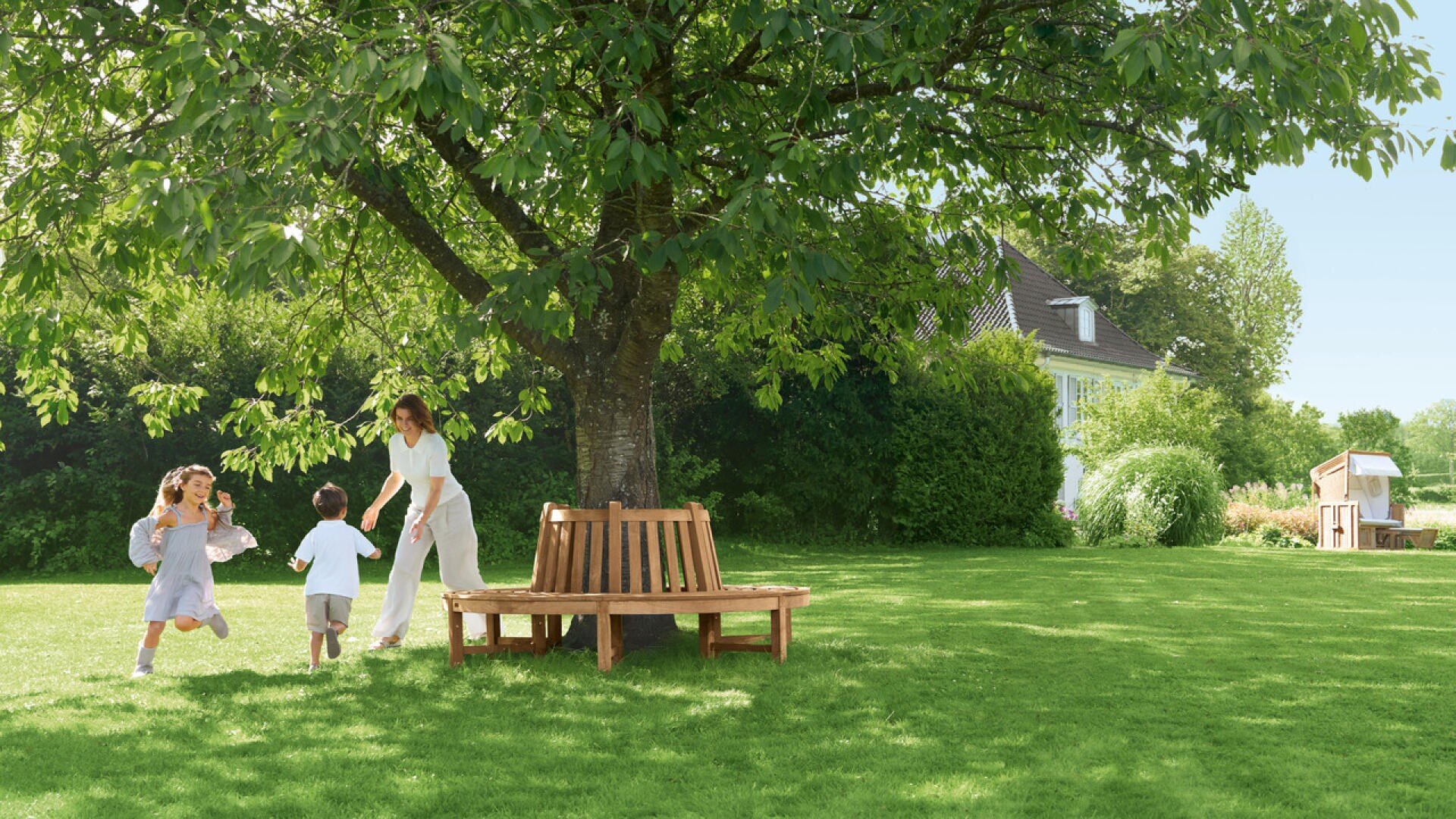 A woman and two children are playing on a sunny, green lawn near a large tree with a round wooden bench. A house and a garden shed can be seen in the background on a clear day.