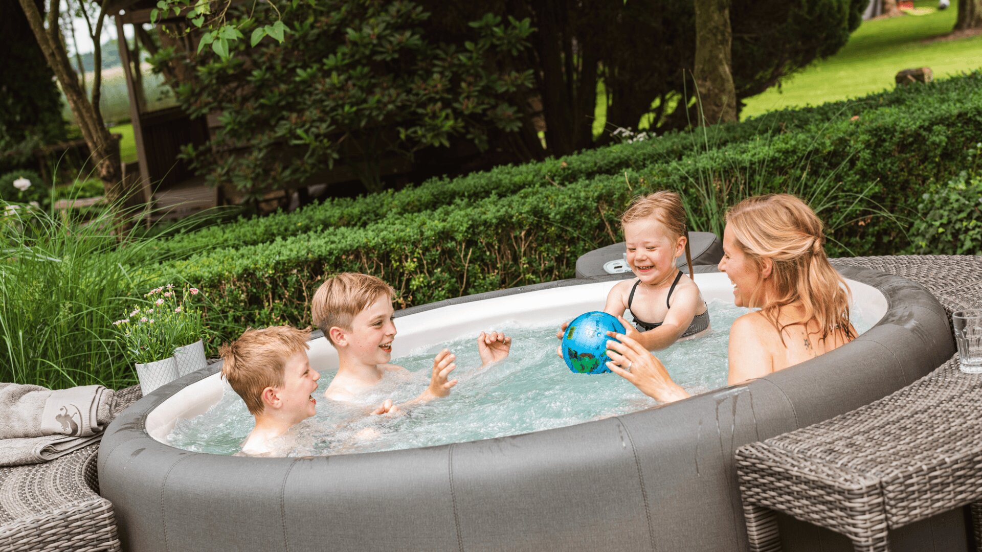 Eine Frau und drei Kinder spielen mit einem kleinen Ball in einem aufblasbaren Whirlpool im Freien, umgeben von Grünpflanzen und Korbmöbeln an einem sonnigen Tag.