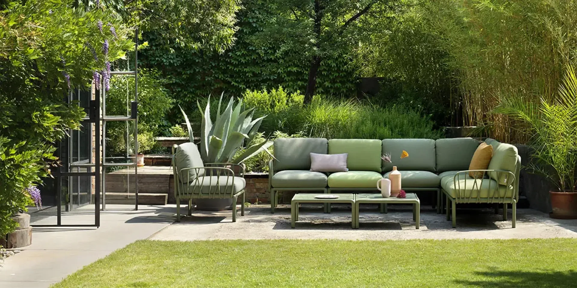 Außenterrasse mit grünem Polstersofa, Sesseln und einem niedrigen Tisch mit Vasen. Umgeben von üppigen grünen Pflanzen und Bäumen, sitzt der Sitzbereich auf einer Terrasse neben einer Glastür und schafft eine entspannte Gartenatmosphäre.