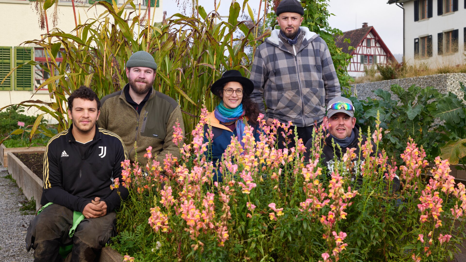 Fünf Personen posieren zusammen in einem Garten mit orangefarbenen und rosafarbenen Blumen im Vordergrund. Drei Männer stehen hinten, zwei Personen sitzen vorne - eine Frau mit Brille und Hut und ein Mann in einer Juventus-Jacke. Im Hintergrund sind Gebäude zu sehen.