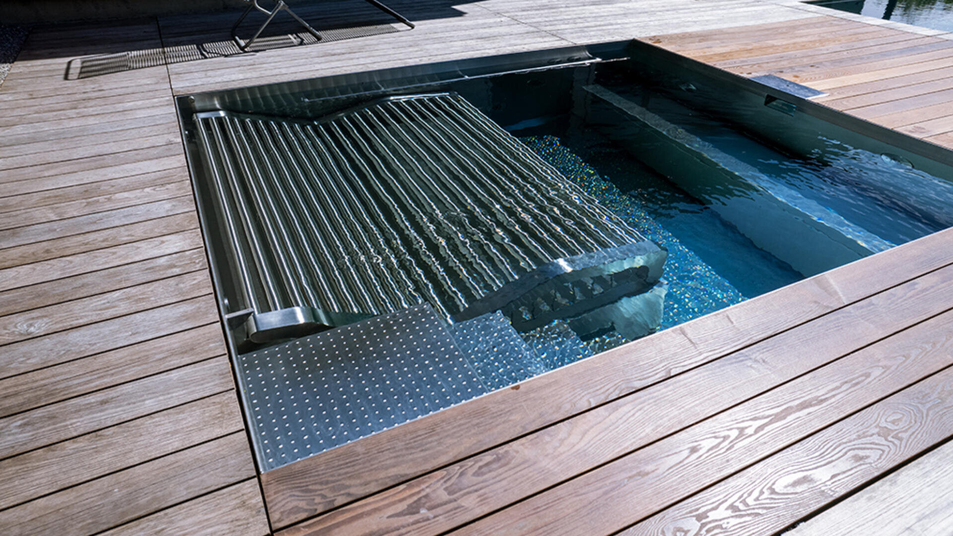Ein moderner, rechteckiger Whirlpool mit einer teilweise angehobenen Abdeckung aus Metalllatten, umgeben von einer Holzterrasse und klarem Wasser im Inneren.