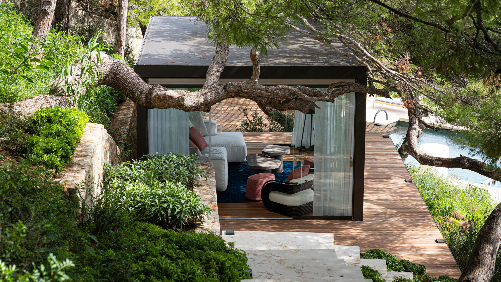 Eine moderne Outdoor-Lounge mit weißen Sofas und einem Pavillon liegt inmitten von üppigem Grün und Bäumen und bietet einen Blick auf den Swimmingpool darunter.