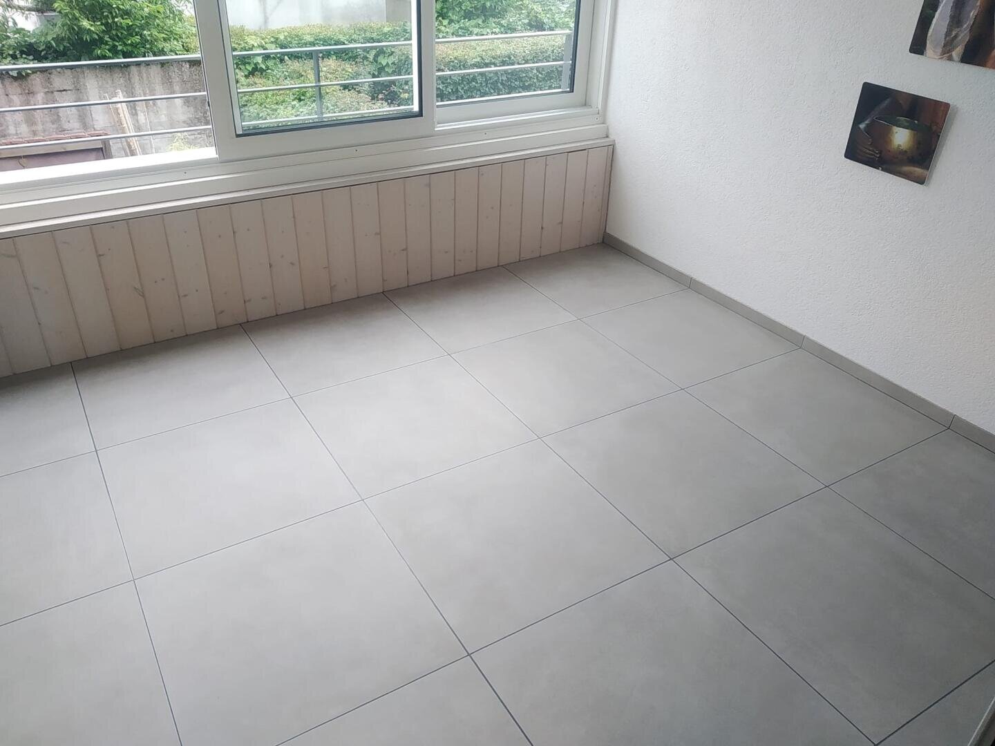 Ein heller Raum mit großen quadratischen grauen Bodenfliesen, einer weißen Wand, einem Fenster mit einem Holzsims und zwei gerahmten Bildern an der Wand. Vor dem Fenster ist Grünzeug zu sehen.