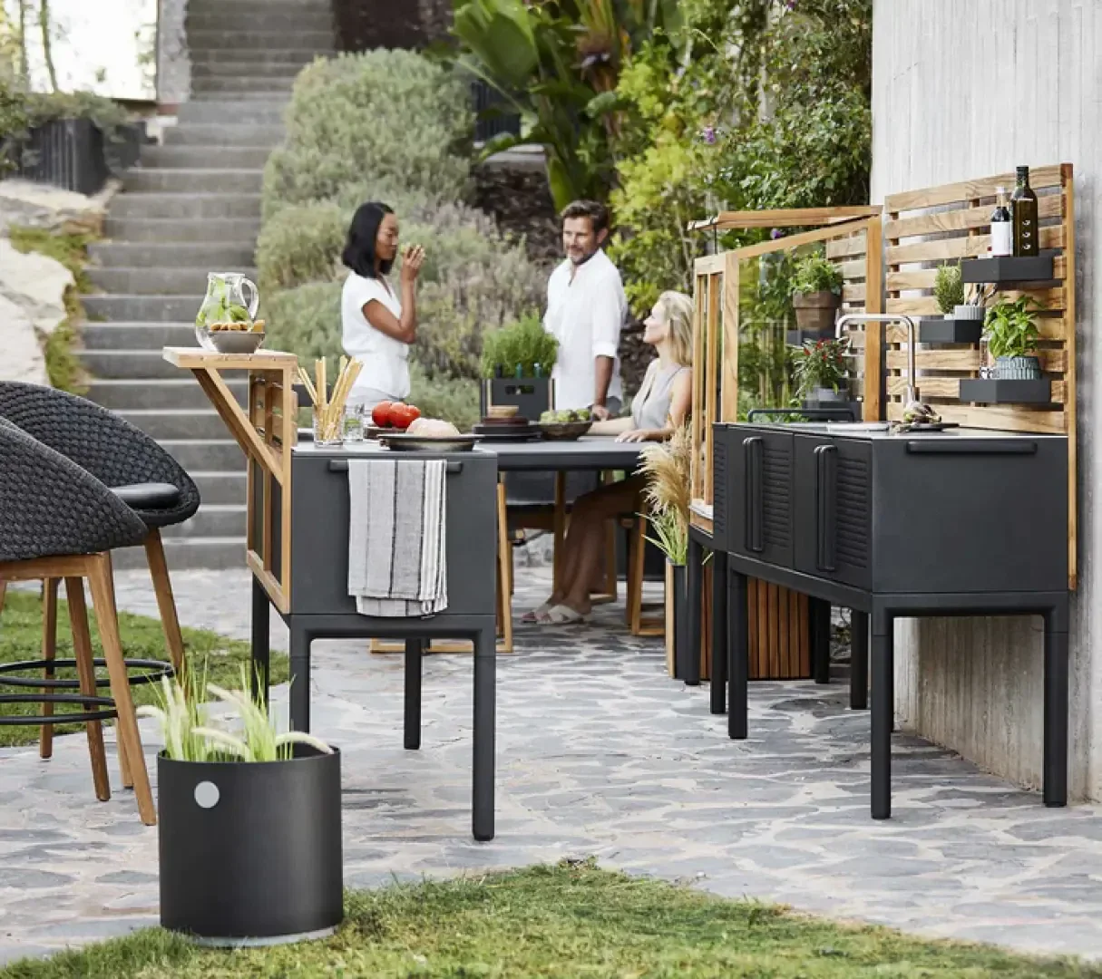 Three people enjoy a conversation in a modern outdoor kitchen and dining area, surrounded by greenery and stylish black furniture on a stone patio.