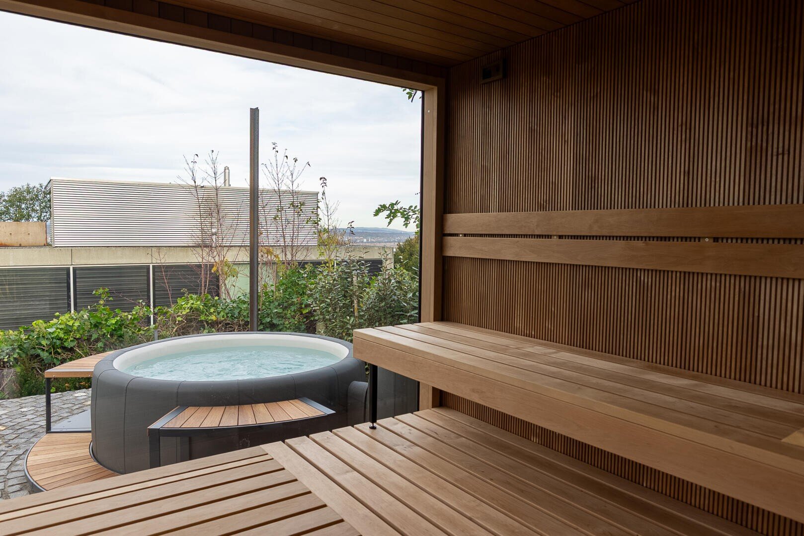 Der Innenraum der Holzsauna mit den gestuften Bänken blickt auf einen Garten mit einem runden Whirlpool, der von Grün umgeben ist, und auf eine Terrasse, die durch ein großes offenes Fenster zu sehen ist.