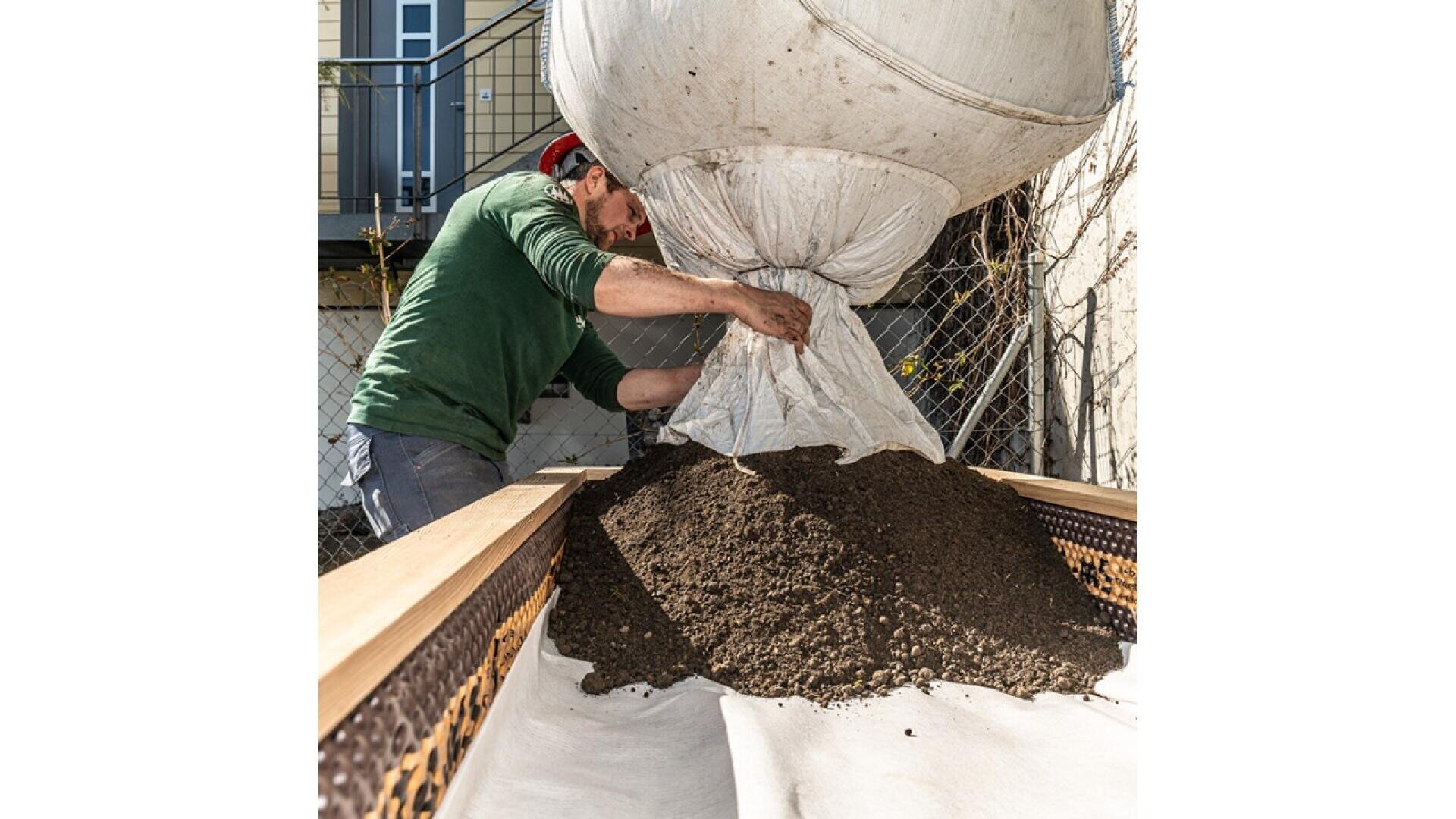 A person in a green shirt and red cap empties a large white sack of soil onto a wooden frame covered with fabric in a backyard area, with a fence and building visible in the background.