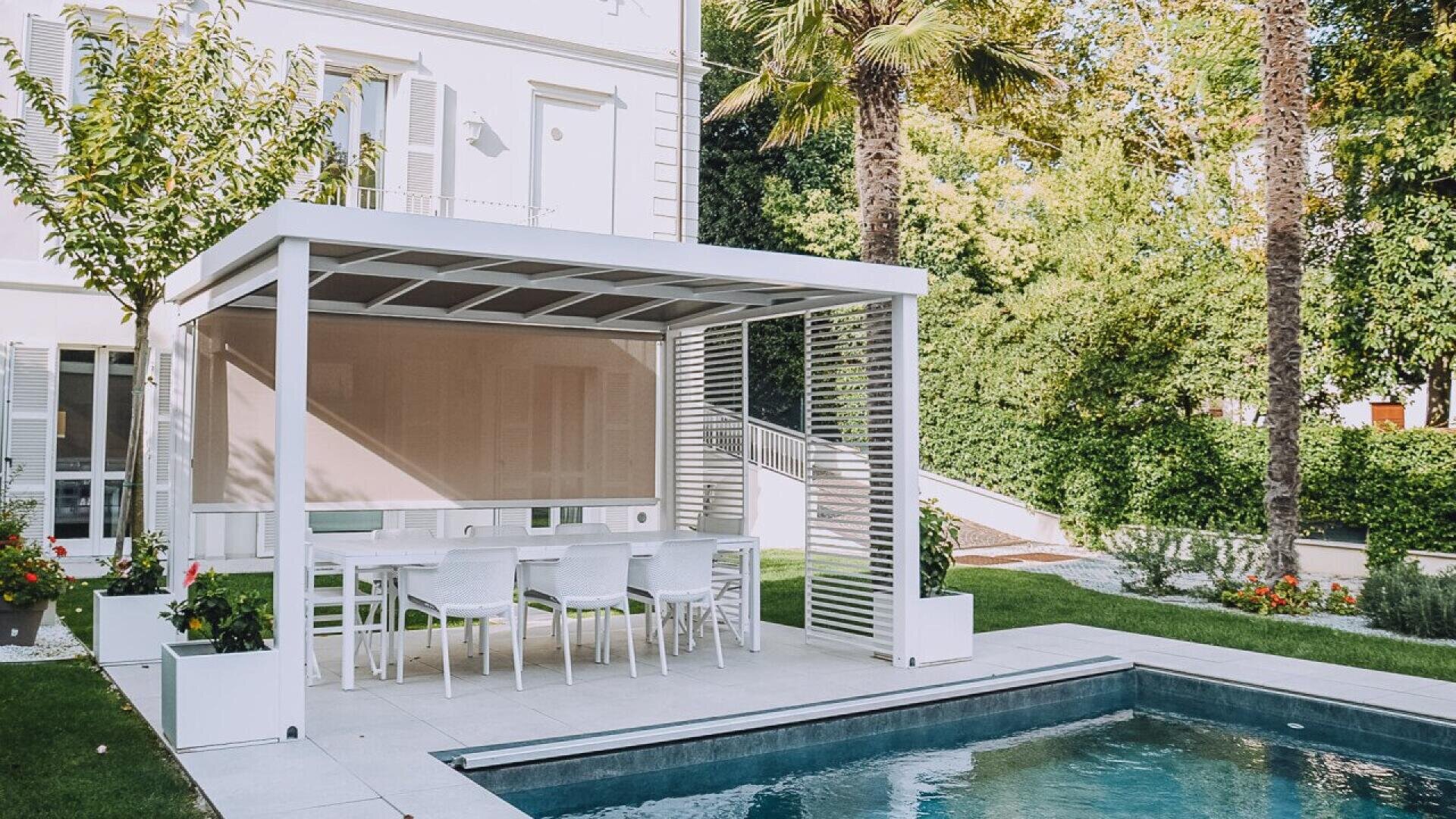 A modern outdoor dining area with white furniture is sheltered by a white pergola beside a swimming pool, surrounded by greenery and adjacent to a white house.