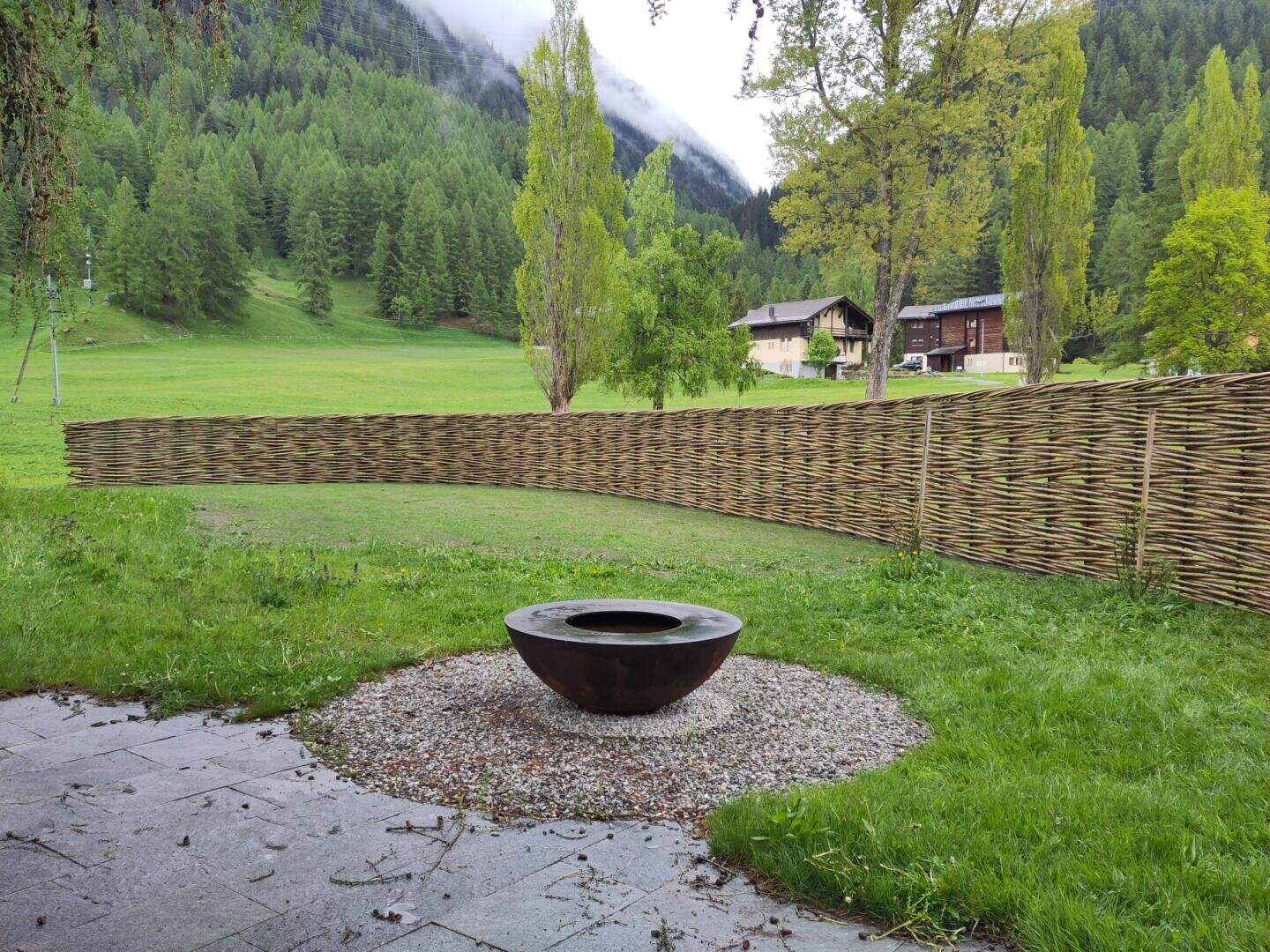 Eine runde Feuerstelle aus Metall liegt auf Kies in einem grasbewachsenen Hof, der von einem geflochtenen Holzzaun begrenzt wird. Hinter dem Zaun erstrecken sich grüne Felder, Bäume und Gebäude, im Hintergrund sind bewaldete Berge zu sehen.
