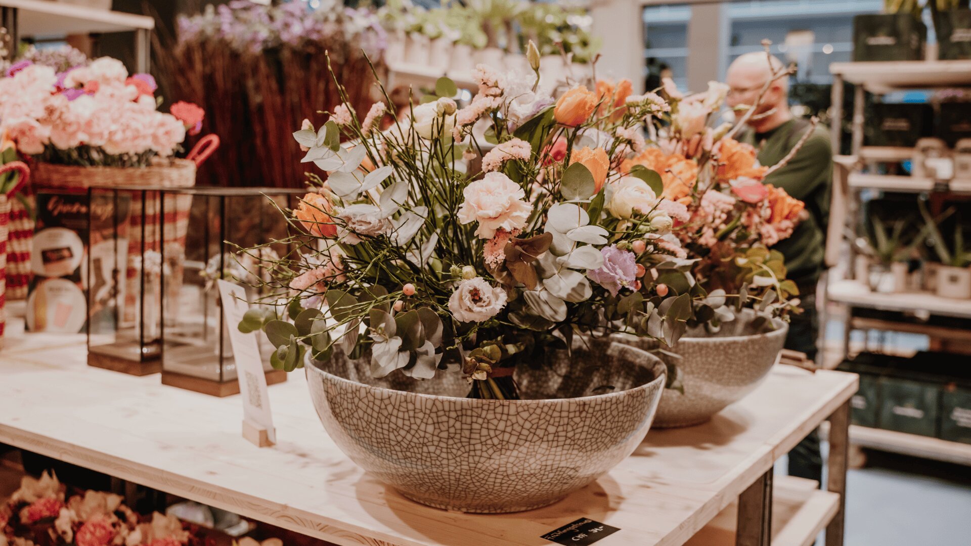 Eine große Keramikschale, gefüllt mit einem Arrangement aus bunten Blumen und Grünzeug, steht auf einem Holztisch in einem hellen, modernen Blumenladen. Im Hintergrund sind weitere Pflanzen und Blumenarrangements zu sehen.