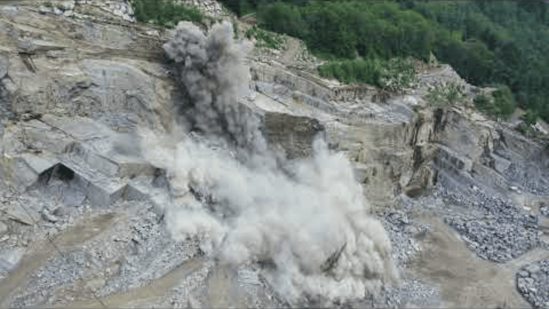 Eine große Explosion wirbelt in einem Steinbruch Staub und Trümmer in die Luft, während im Hintergrund freiliegende Steinwände und Grünflächen zu sehen sind.