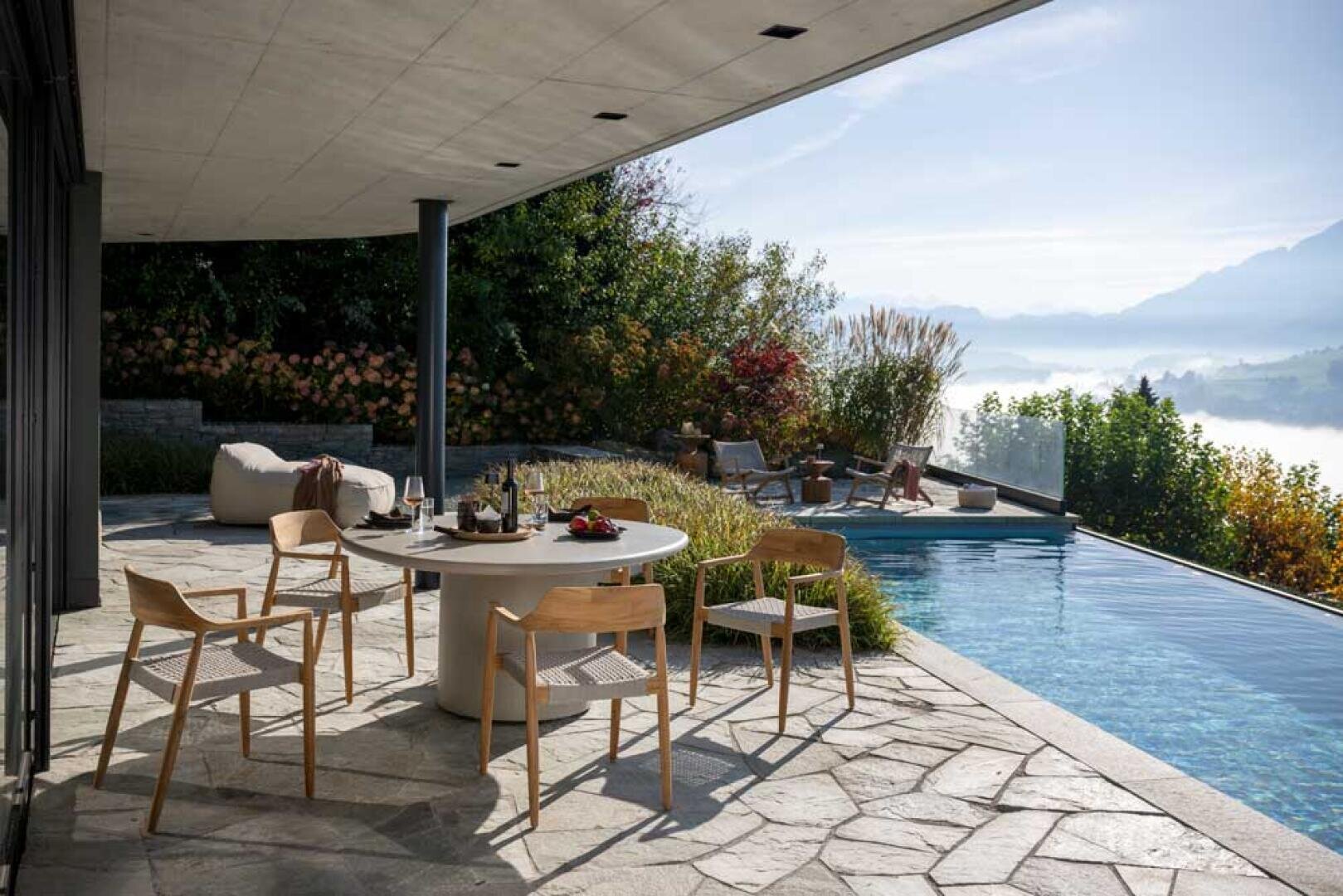 Eine moderne Terrasse mit rundem Esstisch und Holzstühlen befindet sich neben einem Infinity-Pool mit Blick auf ein nebliges Tal und die Berge, während sich zwei Personen auf Liegen im Hintergrund entspannen.