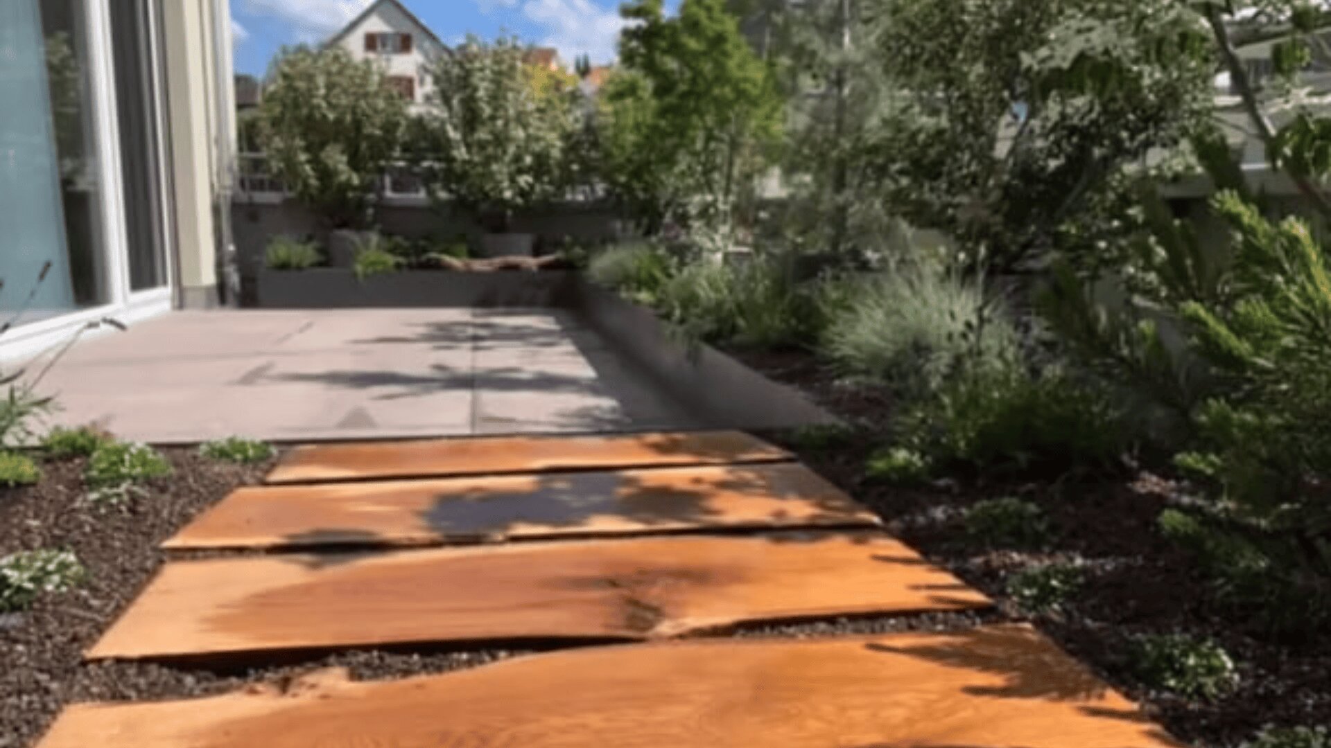 Ein Gartenweg aus breiten Holzbohlen führt durch eine Grünanlage mit Pflanzen und Sträuchern, neben einem modernen Gebäude mit großen Fenstern und einem gepflasterten Innenhof im Hintergrund.
