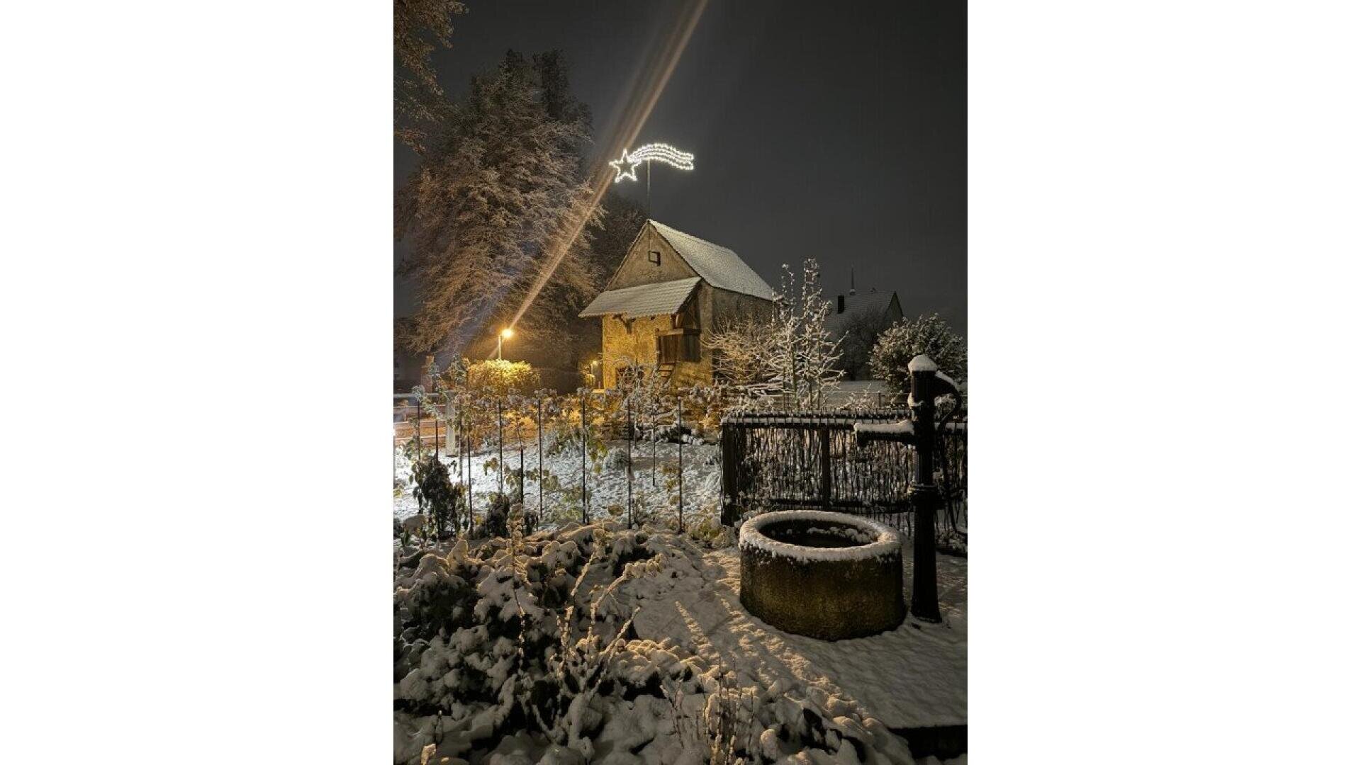 A small house with a steep roof is surrounded by a snowy yard and trees at night. Warm lights illuminate the scene, and a glowing star-shaped decoration hangs above the house. A stone well is in the foreground.