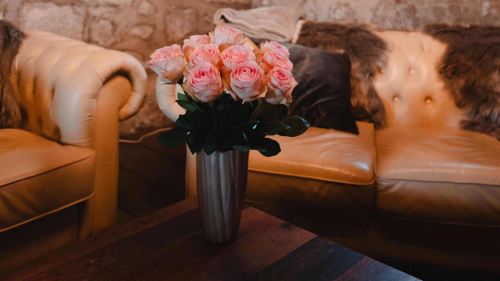 Ein Strauß rosafarbener Rosen in einer silbernen Vase steht auf einem hölzernen Couchtisch zwischen zwei beigen Ledersofas mit Fellüberwürfen vor einem Hintergrund aus Stein.