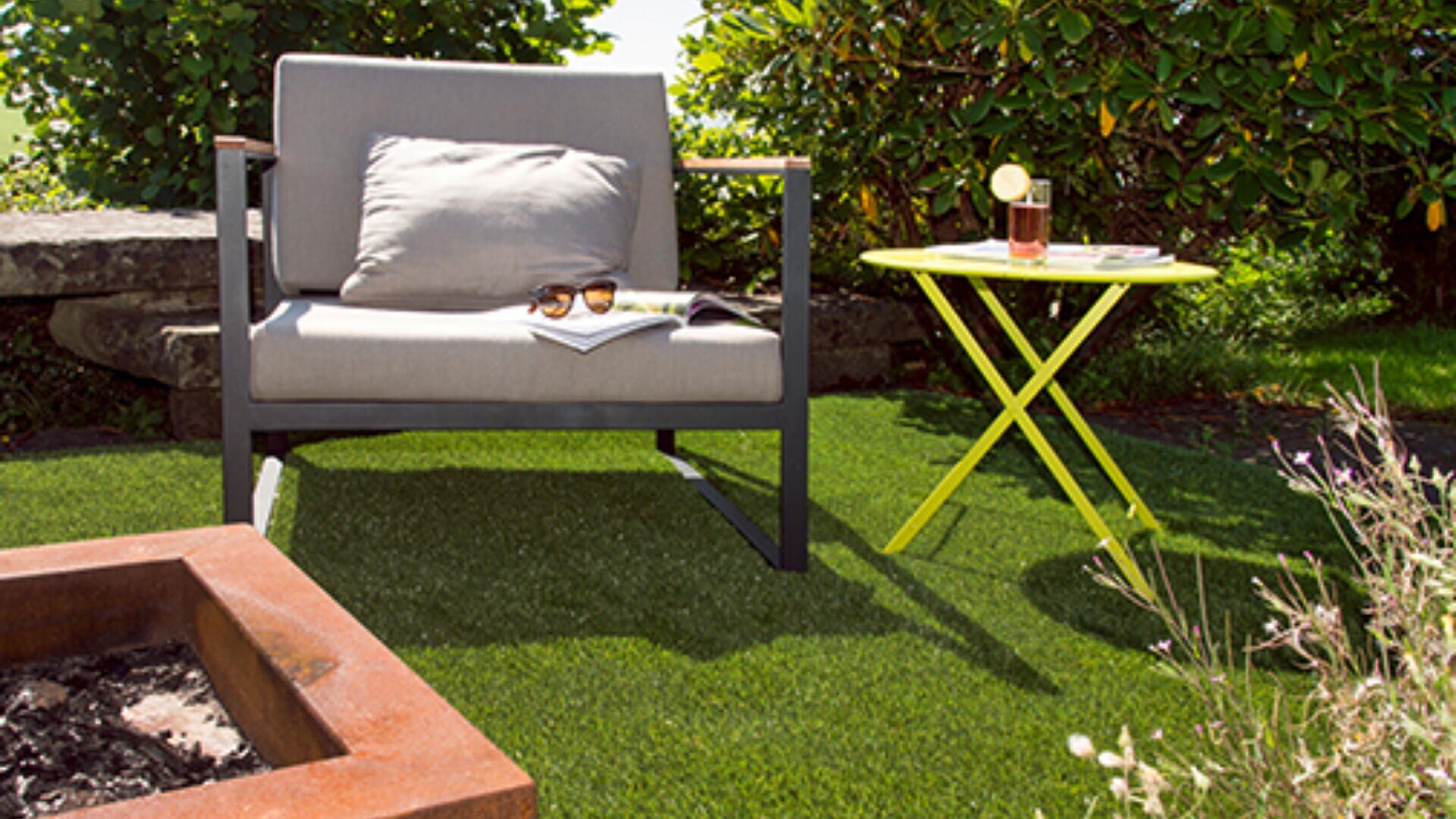 Ein moderner Outdoor-Stuhl mit einem Kissen sitzt auf grünem Gras neben einem kleinen gelben Tisch mit einem Getränk und einem Buch. Auf dem Arm des Stuhls liegen eine Sonnenbrille und eine Zeitschrift, im Vordergrund ist eine Feuerstelle zu sehen.