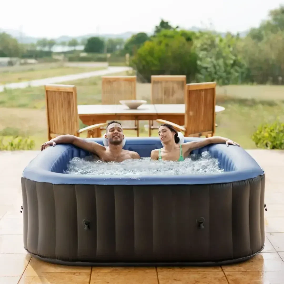 Ein Mann und eine Frau entspannen sich in einem aufblasbaren Whirlpool auf einer Terrasse, lächelnd und mit zurückgeneigtem Kopf, umgeben von sprudelndem Wasser. Im Hintergrund sind Gartenmöbel und Bäume zu sehen.