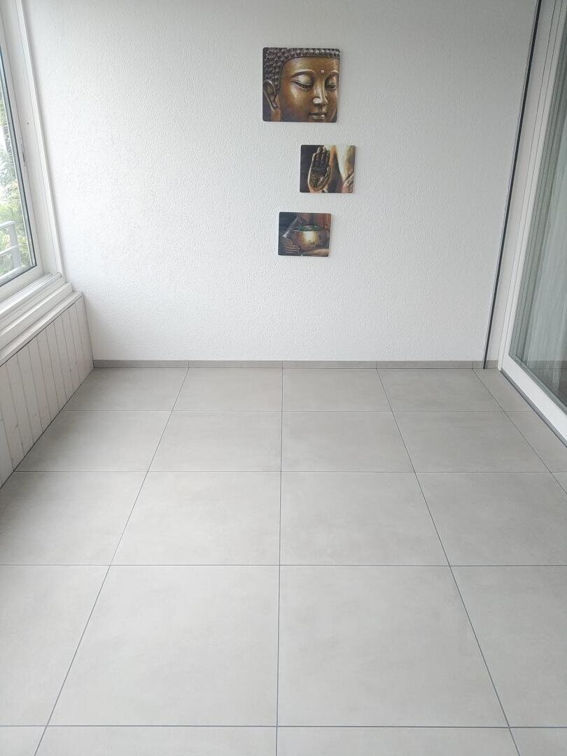 Ein minimalistischer Raum mit hellgrauem Fliesenboden, weißen Wänden und drei Kunstwerken zum Thema Buddha, die in einer vertikalen Linie an der Wand hängen. Es gibt große Fenster und eine Glastür auf der rechten Seite.