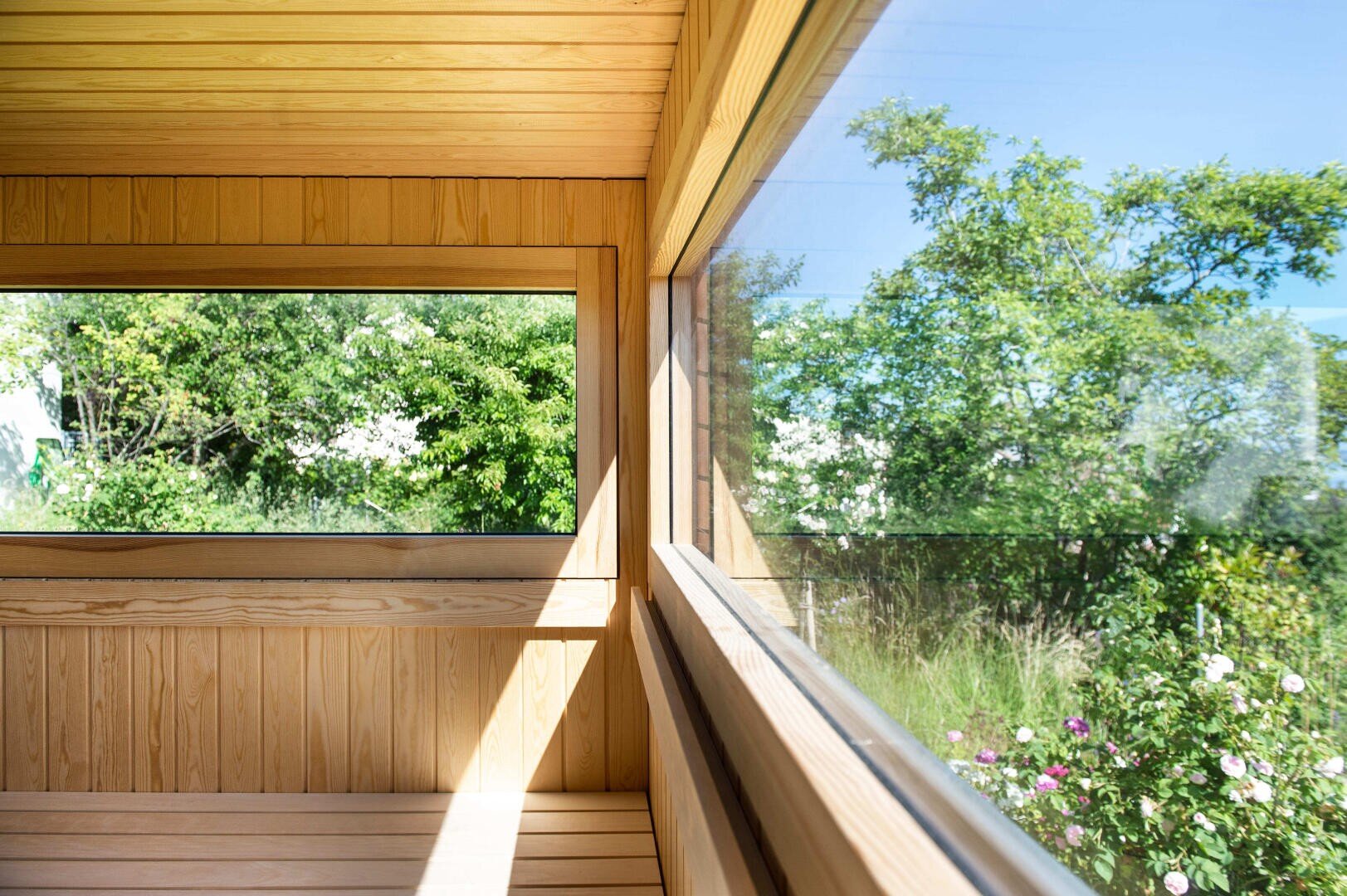 A bright, wooden sauna cabin with large windows offers a view of a lush, green garden with trees and flowering shrubs under a clear blue sky. Sunlight streams into the room, creating a warm and inviting atmosphere.