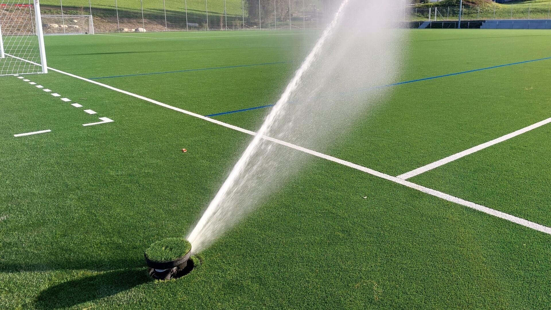 Ein Sprinkler sprüht Wasser diagonal über einen grünen Kunstrasen-Sportplatz, in der Nähe von weißen Begrenzungslinien und einem Fußballtor im Hintergrund.