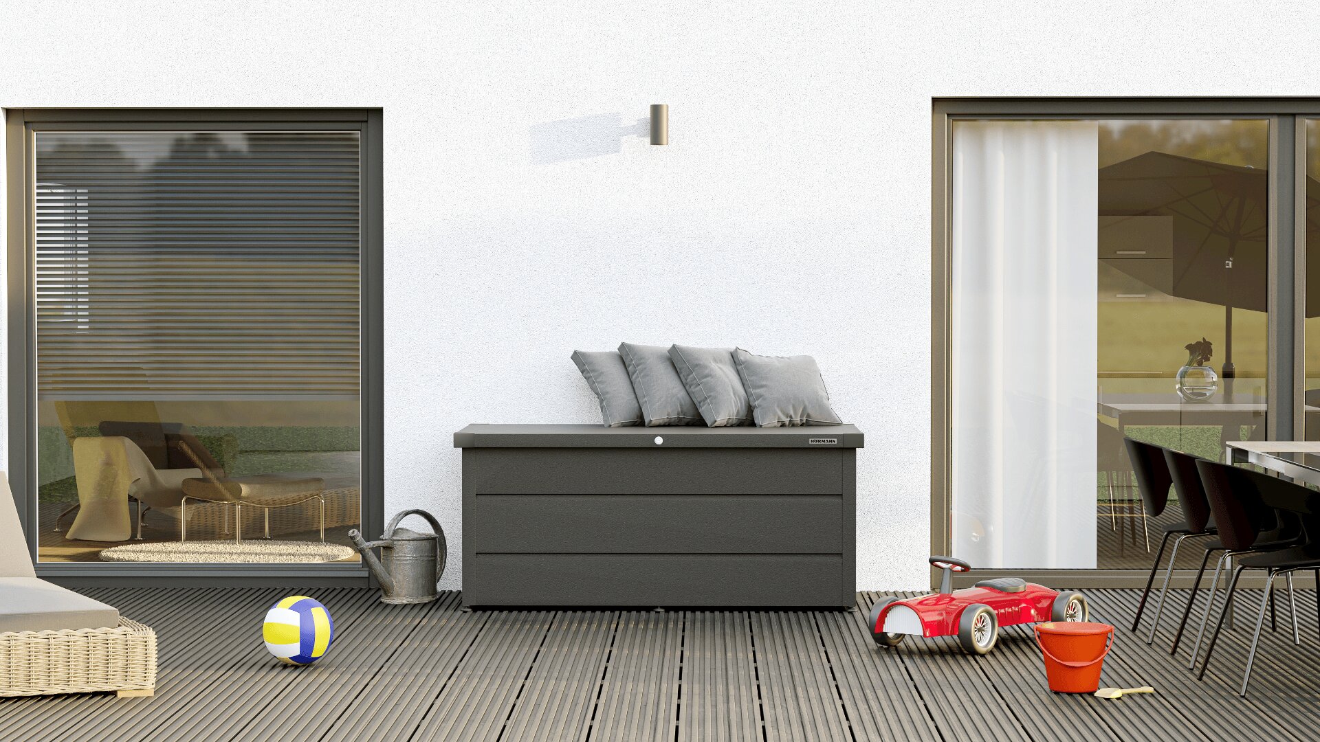 Eine moderne Terrasse mit einer mit grauen Kissen ausgelegten Aufbewahrungsbox, umgeben von einer Gießkanne, einem Strandball, einem Spielzeugauto, einem roten Eimer und einer kleinen Schaufel. Schiebetüren führen zum Wohn- und Essbereich im Inneren.