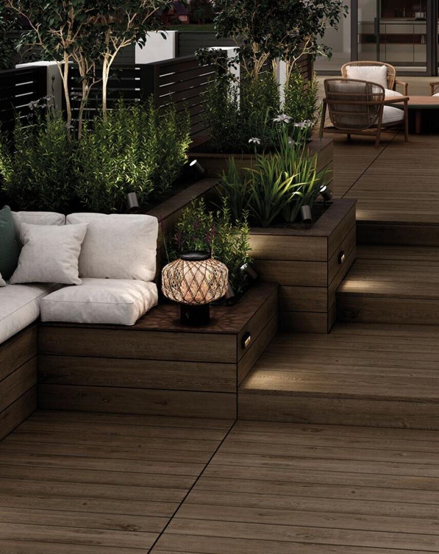 Moderne Außenterrasse mit Holzterrasse, eingebauten Sitzbänken mit weißen Kissen, mit Grünzeug gefüllten Pflanzkästen, stimmungsvoller Beleuchtung und einer Weidenlaterne auf einem Beistelltisch. Im Hintergrund sind gemütliche Stühle zu sehen.
