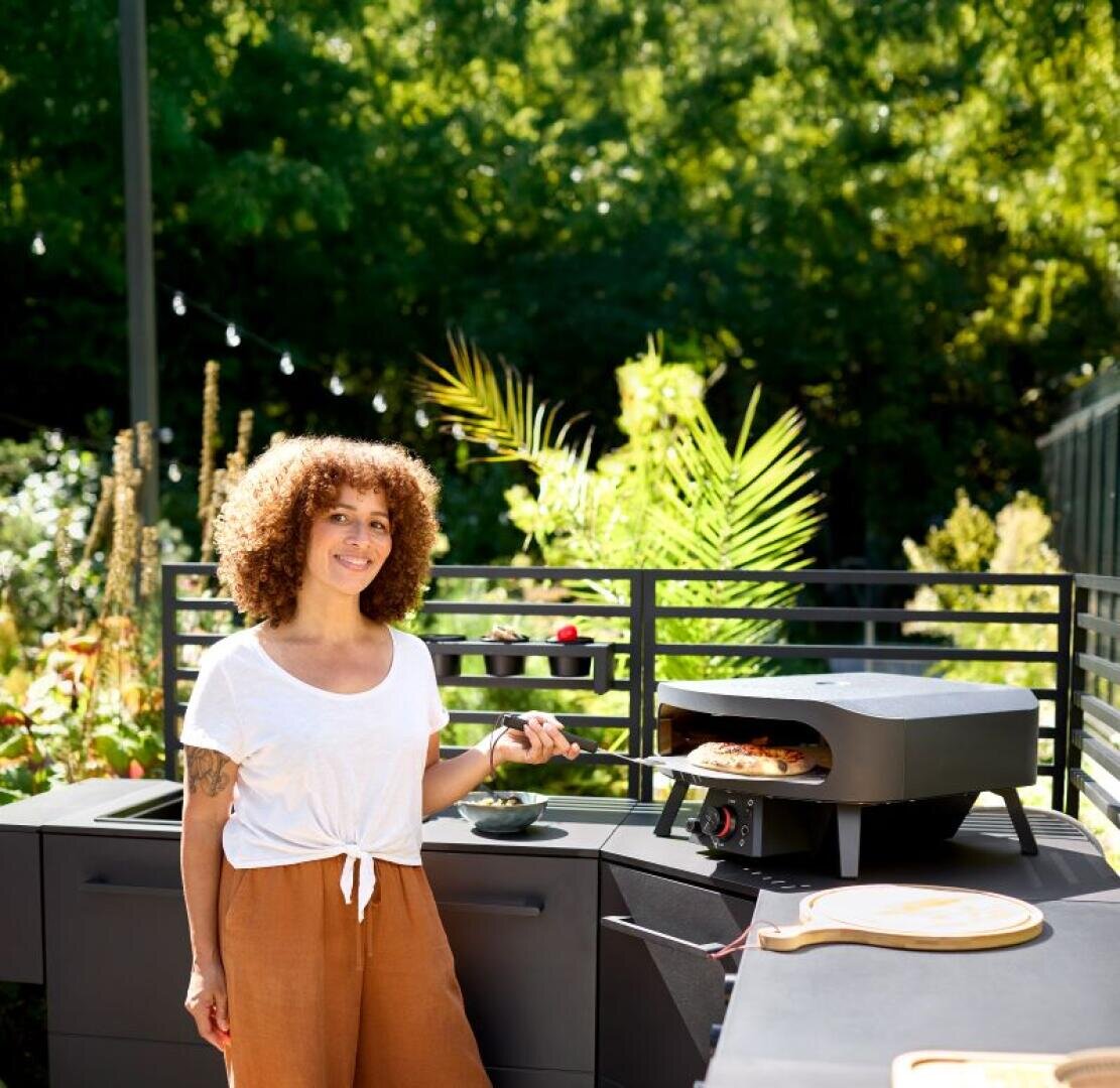 Eine Frau mit lockigem Haar, die ein weißes Hemd und eine braune Hose trägt, lächelt, während sie im Freien neben einem schwarzen Pizzaofen mit einer Pizza darin steht. Im sonnigen Hintergrund sind grüne Pflanzen und Bäume zu sehen.