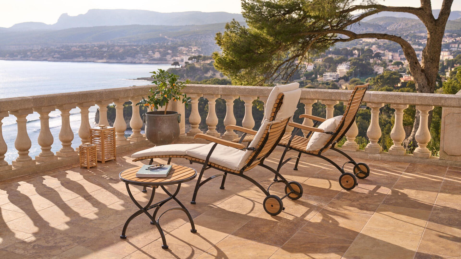 Zwei gepolsterte Holzsessel und ein runder Beistelltisch stehen auf einer sonnigen Terrasse mit Blick auf das Meer, mit einer Topfpflanze in der Nähe und entfernten Hügeln und Gebäuden im Hintergrund.