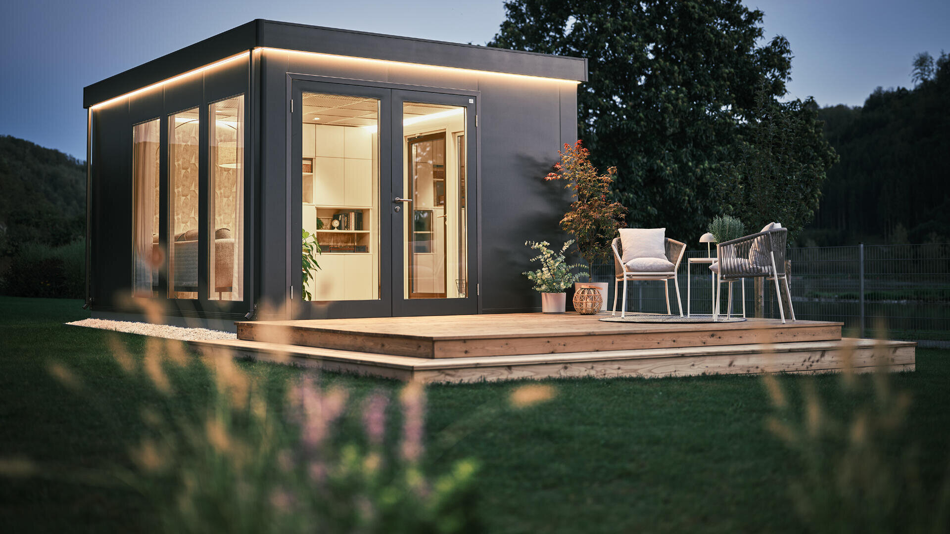 Ein modernes Gartenstudio mit großen Fenstern befindet sich auf einer Holzterrasse mit zwei Stühlen, einem kleinen Tisch und Topfpflanzen, umgeben von Gras und Bäumen in der Abenddämmerung im Freien.