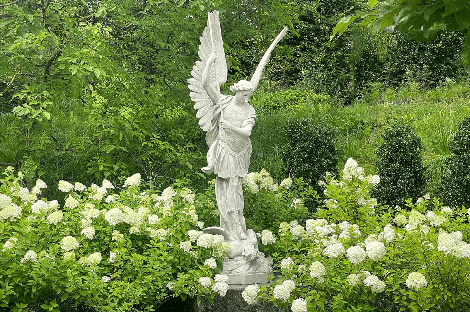 Eine weiße Steinstatue eines Engels mit Flügeln steht inmitten blühender weißer Blumen und üppiger grüner Sträucher in einem Garten. Der Engel hält ein Schwert in der Hand und blickt in die Ferne. Der Hintergrund ist mit Grünzeug gefüllt.