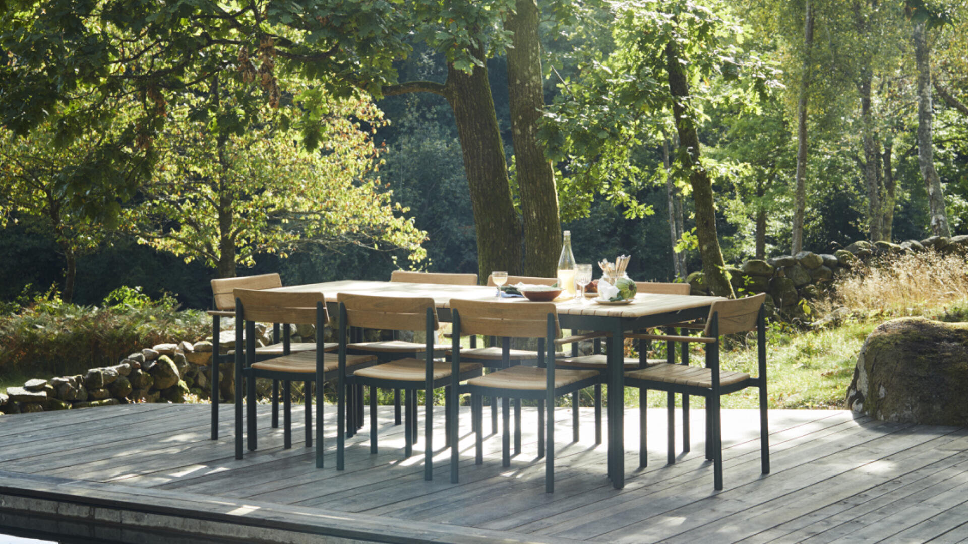 Ein hölzerner Esstisch mit sechs passenden Stühlen steht auf einer Terrasse, umgeben von üppigen grünen Bäumen und Sonnenlicht, auf dem Tisch stehen Gläser, eine Flasche und Lebensmittel.