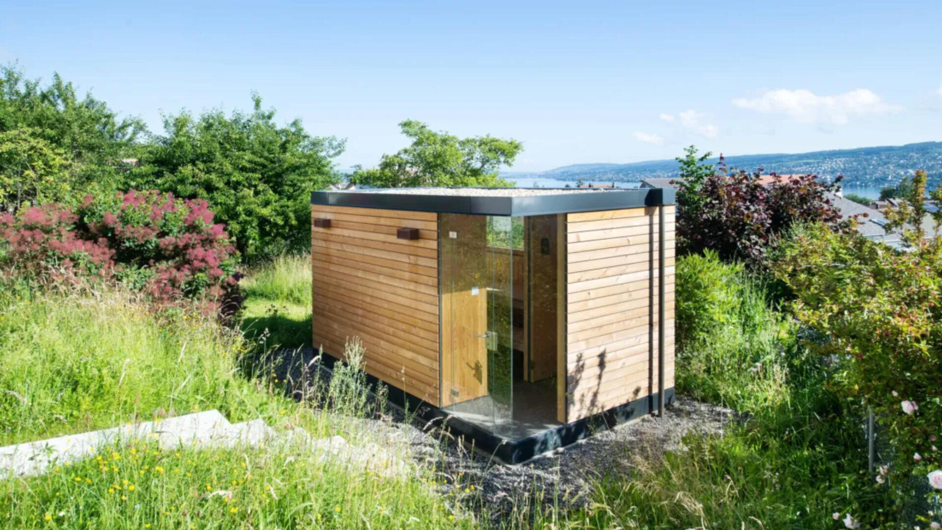 Eine kleine, moderne Holzhütte mit großen Glasfenstern ist von hohem Gras, Büschen und Bäumen umgeben und bietet einen Blick auf ein Gewässer im Hintergrund unter einem klaren blauen Himmel.