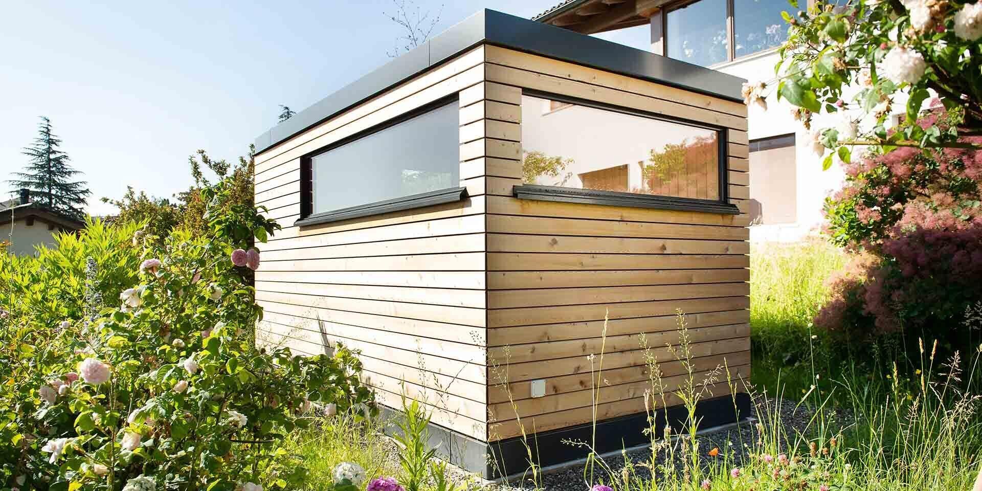 A small, modern wooden shed with horizontal cladding and a large rectangular window stands in a lush, green garden surrounded by various plants and flowers.