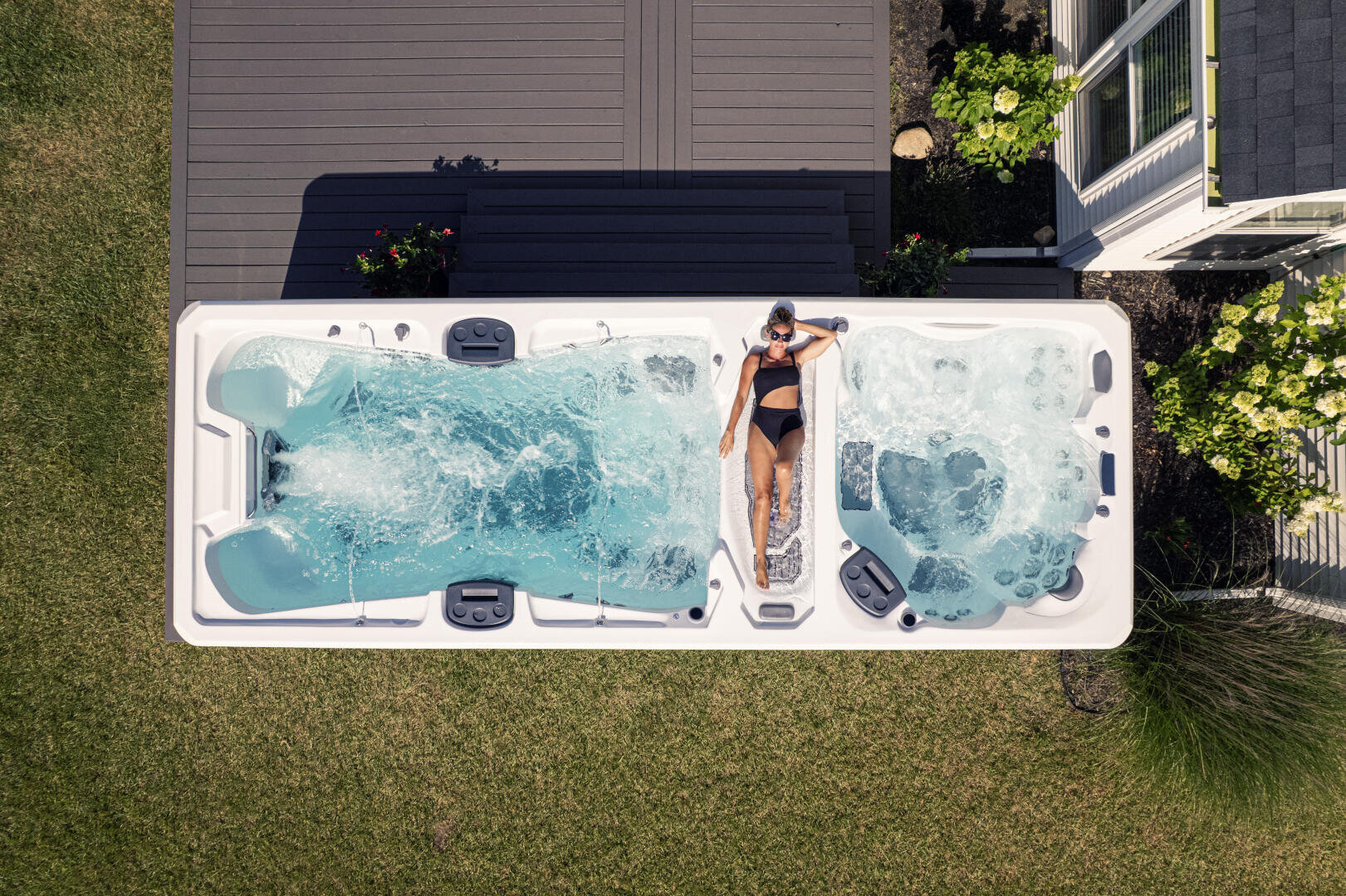Eine Frau in einem schwarzen Badeanzug entspannt sich im Whirlpool eines großen Freibads, das einen Schwimmbereich mit einem Whirlpool kombiniert, neben einer Terrasse und grünem Rasen, von oben gesehen.