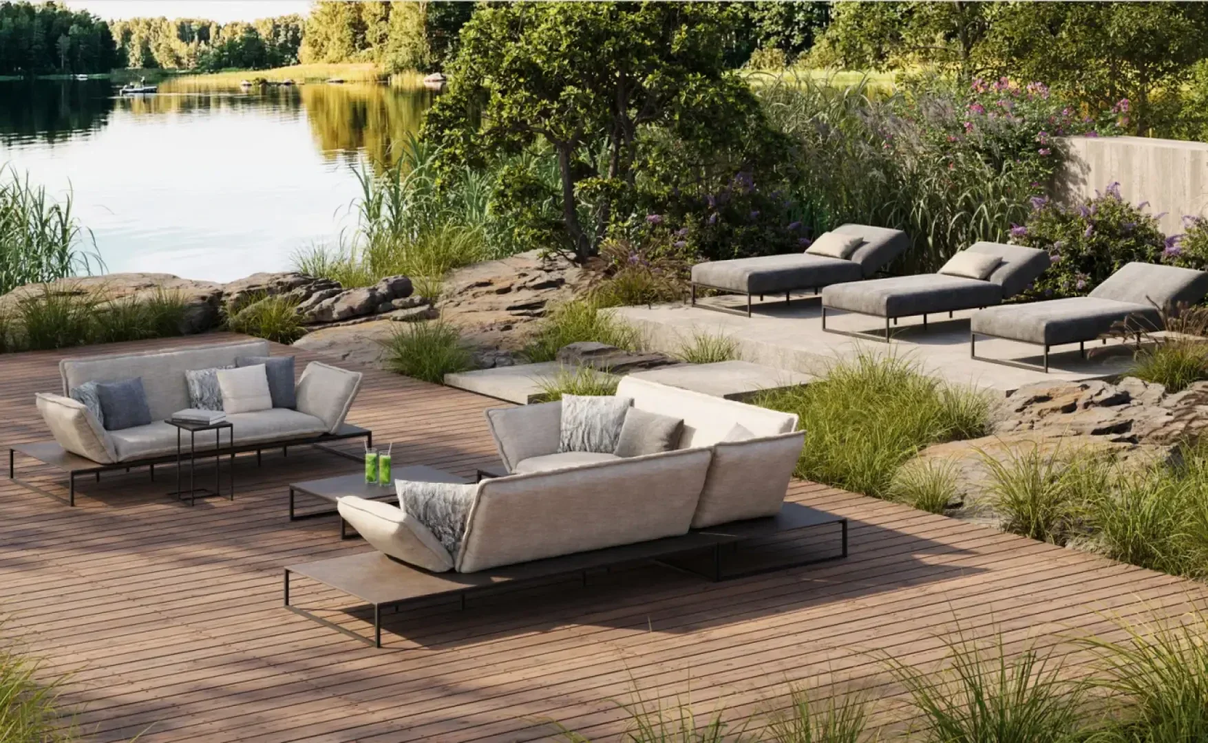 Modern outdoor lounge area with upholstered sofas and armchairs on a wooden deck overlooking a tranquil lake, surrounded by rocks, tall grass, trees and lush greenery.