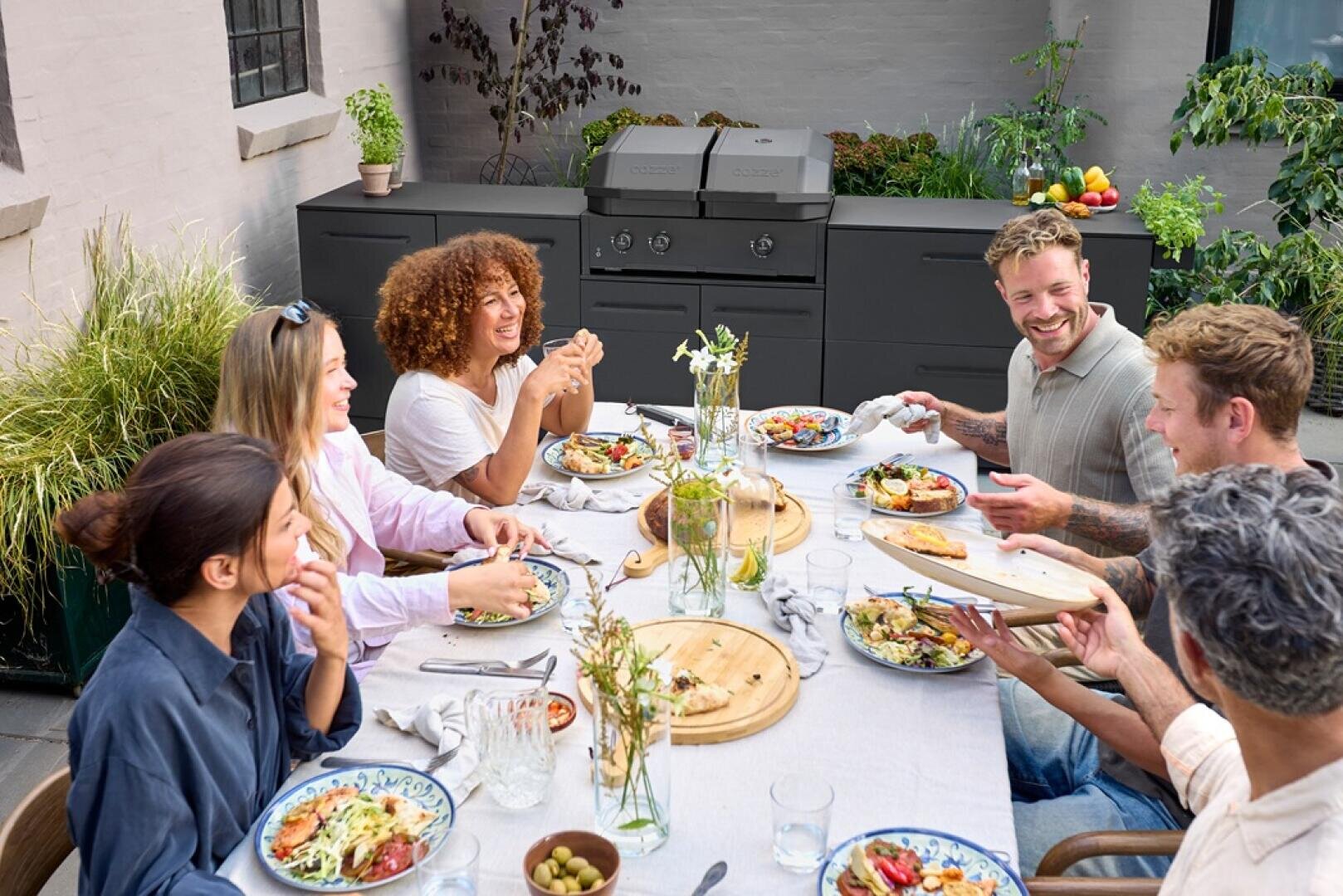 Eine Gruppe von sechs Personen sitzt an einem Tisch im Freien und genießt eine gemeinsame Mahlzeit, lächelt und lacht. Auf dem Tisch stehen Teller mit Essen, Getränken und leerem Geschirr. Im Hintergrund sind ein Grill und Pflanzen zu sehen.