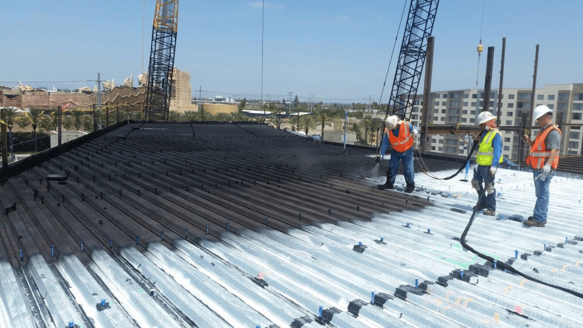 Drei Bauarbeiter in Schutzkleidung stehen auf einem Metallgerüst und arbeiten mit großen Schläuchen. Im Hintergrund sind Kräne und Gebäude unter einem klaren blauen Himmel zu sehen.