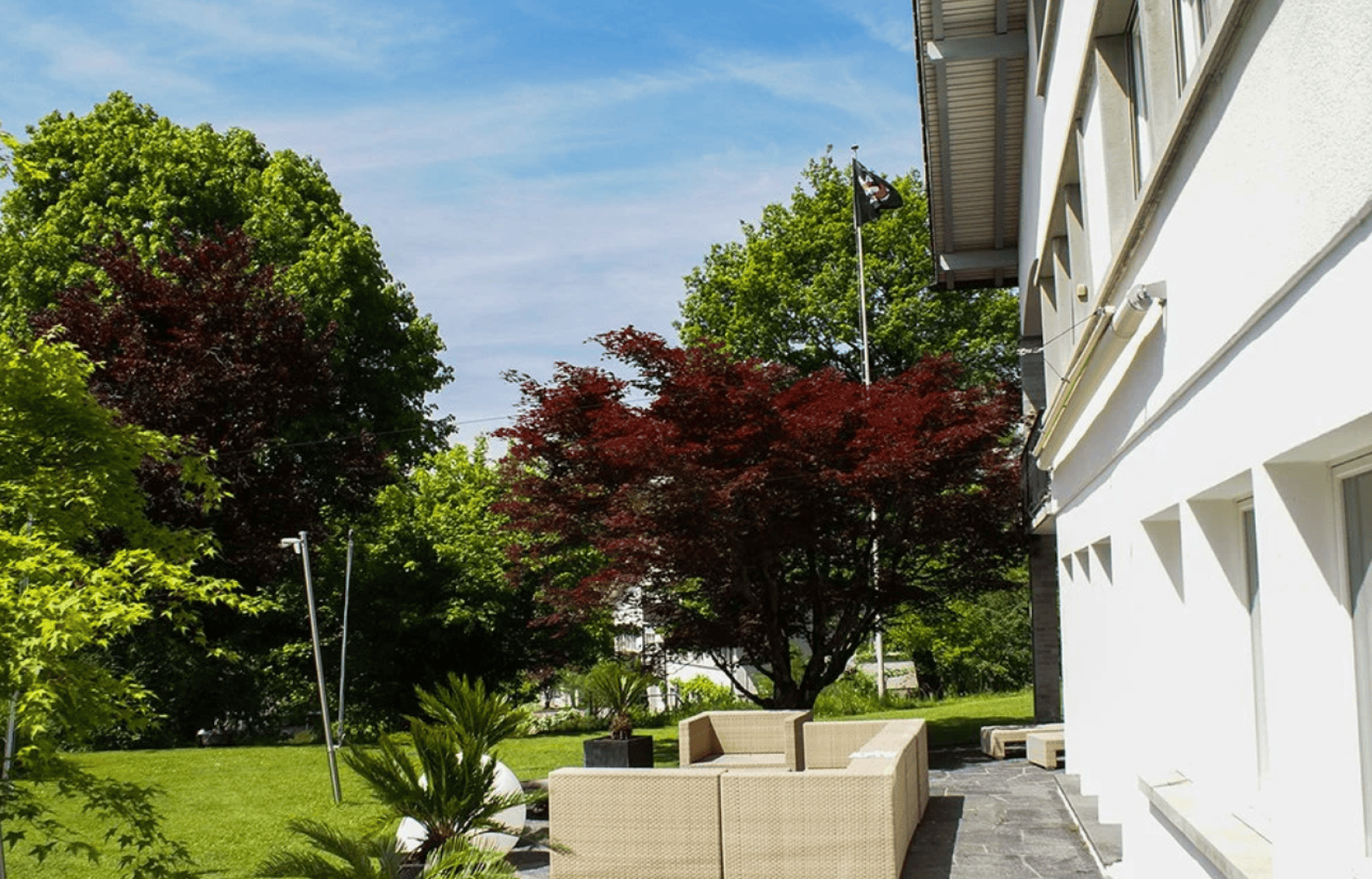 Outdoor patio with beige wicker furniture beside a modern white building, surrounded by lush green grass, leafy trees, and a red-leaved tree under a blue sky with scattered clouds.
