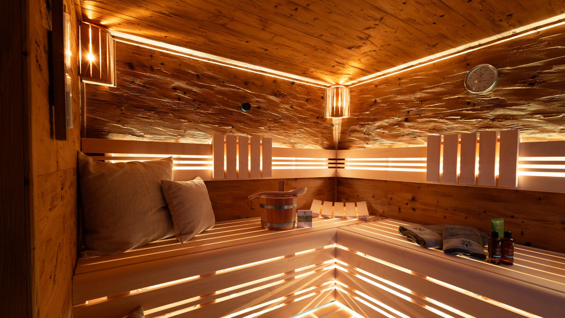 Eine gemütliche Holzsauna mit warmer Beleuchtung, Holzbänken, einem Kissen, einem Eimer mit einer Kelle, ordentlich gefalteten Handtüchern und einer Uhr an der Wand, die eine entspannende und einladende Atmosphäre schafft.