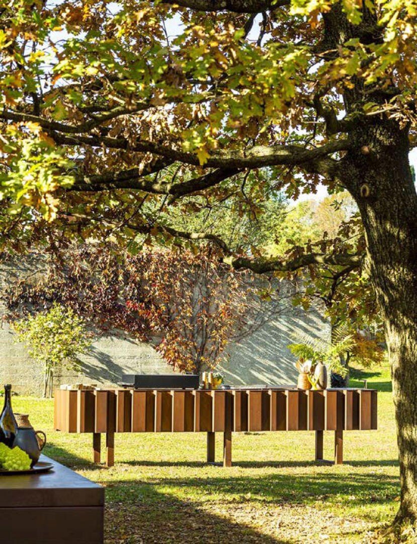 Modern outdoor furniture sits on a sunlit lawn beneath a large tree with green and yellowing leaves, creating a relaxed, natural setting. Decorative items and drinks are arranged on the tables in the background.