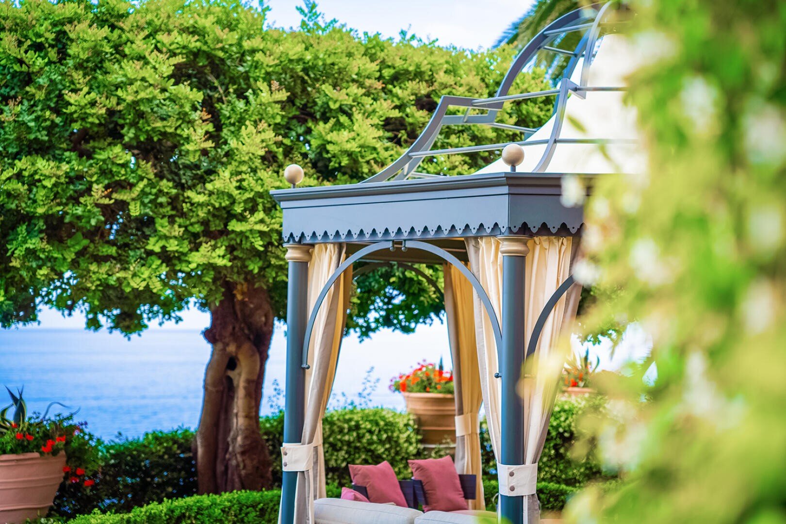 Eleganter Pavillon mit Metallsäulen und dekorativem Dach, umgeben von üppigem Grün und Topfpflanzen. Im Hintergrund sind das Meer und der blaue Himmel zu sehen. Unter der Gartenlaube sind teilweise gepolsterte Sitzgelegenheiten zu sehen.