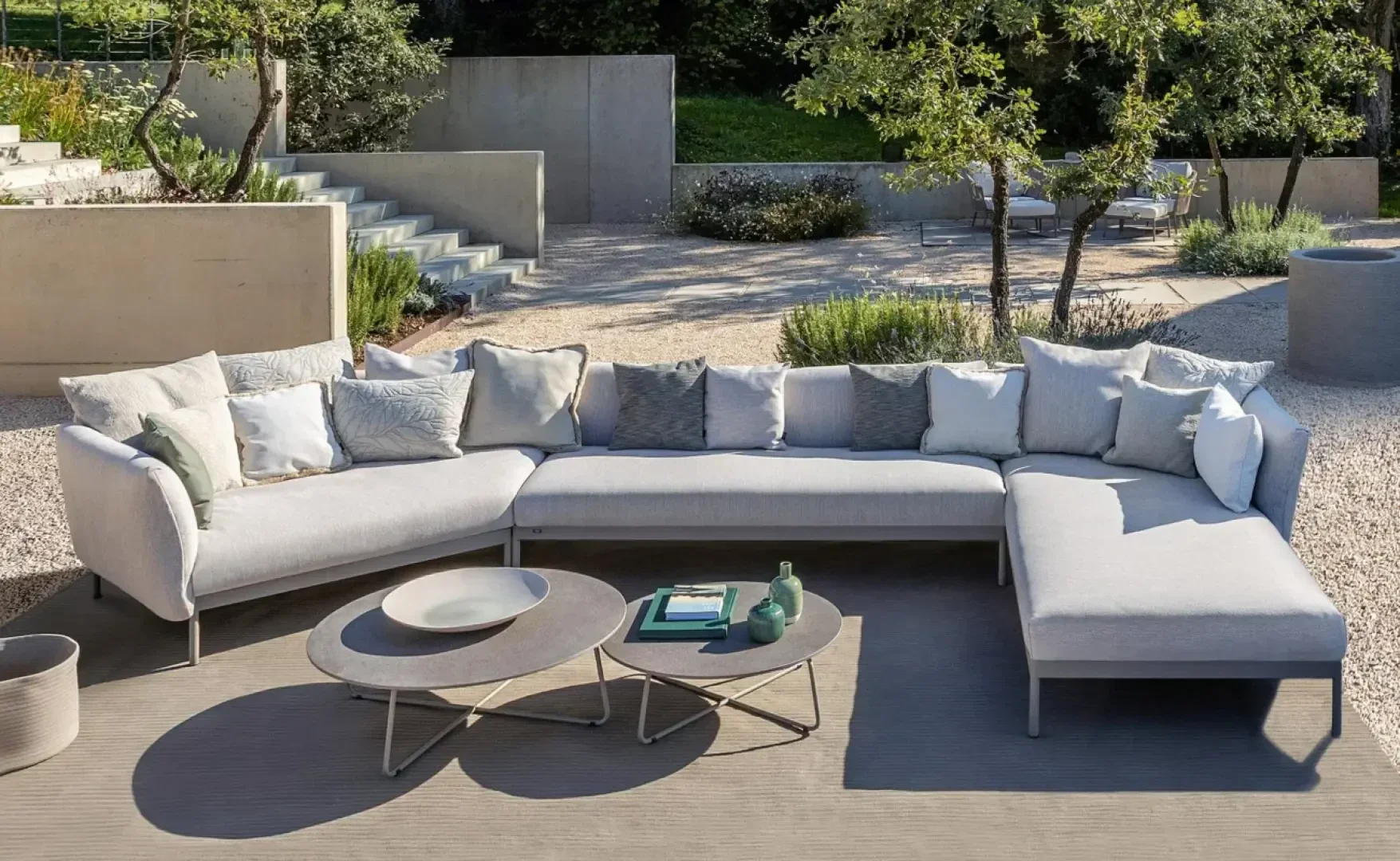 A modern outdoor terrace with a light gray L-shaped sectional sofa, numerous cushions, a coffee table with books, vases and bowls, all on a terrace surrounded by gravel, trees and green plants.