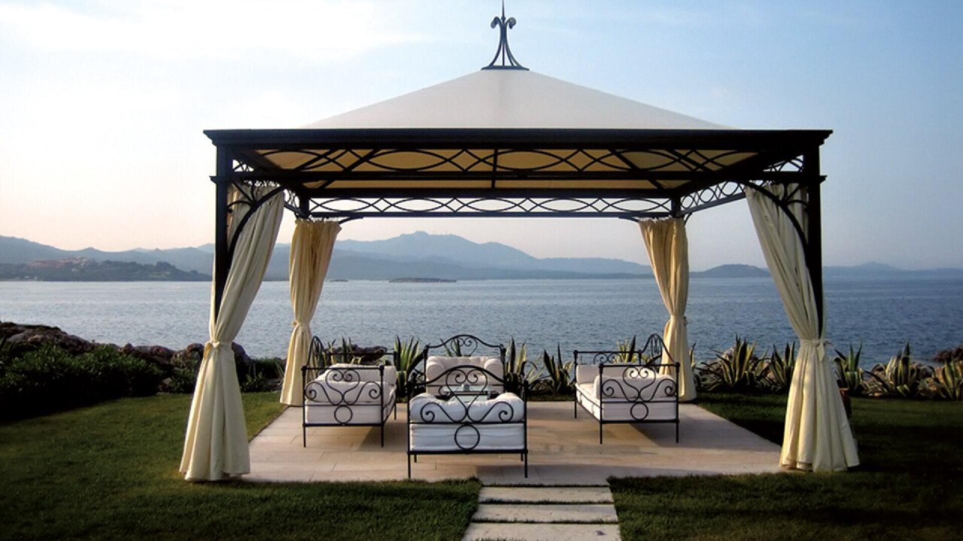 A gazebo with cream curtains and four cushioned metal chairs overlooks a calm body of water and distant hills, surrounded by greenery on a sunny day.